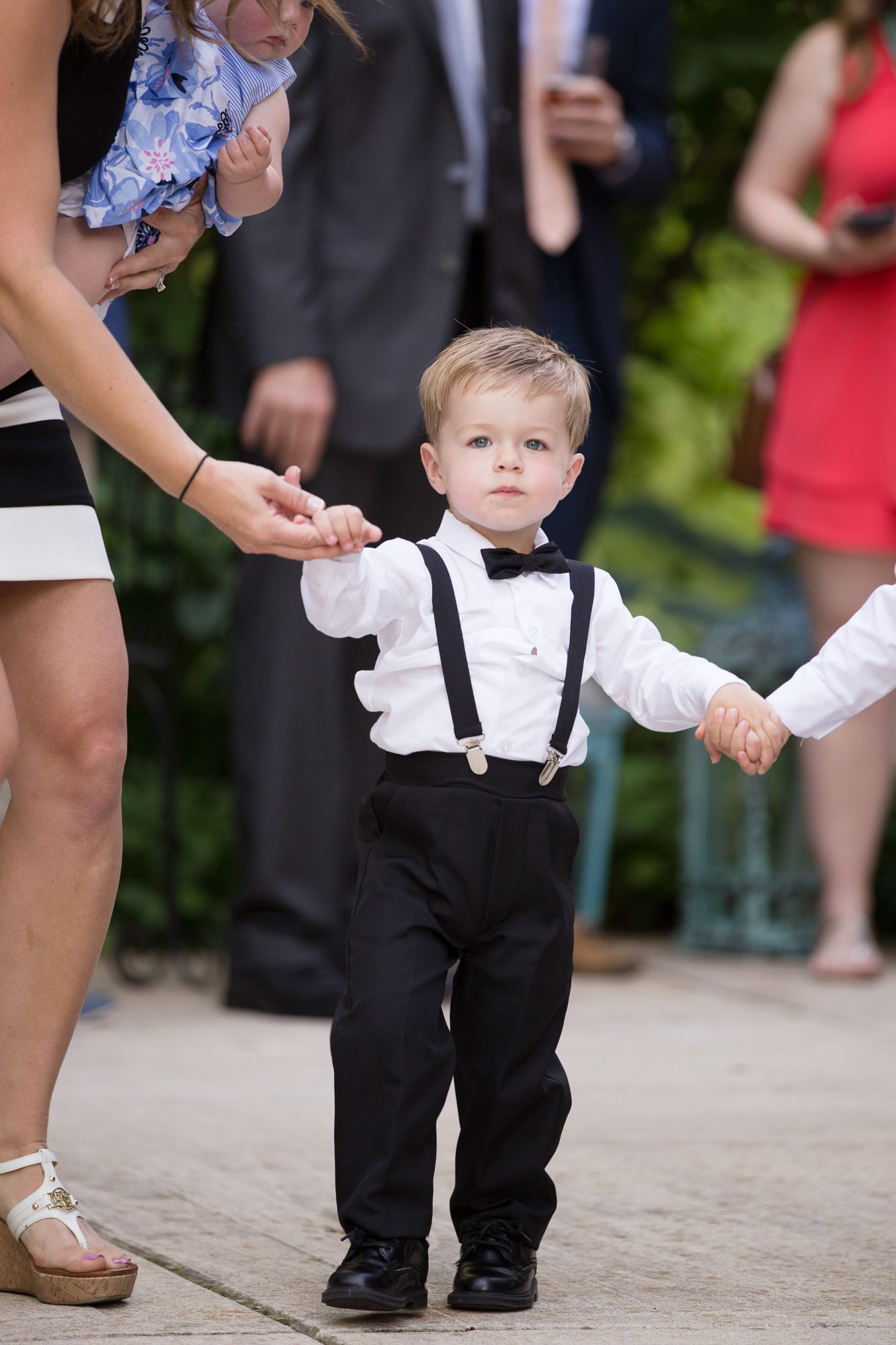A toddler in black suspenders and white shirt takes steps while being supported by adults' hands.