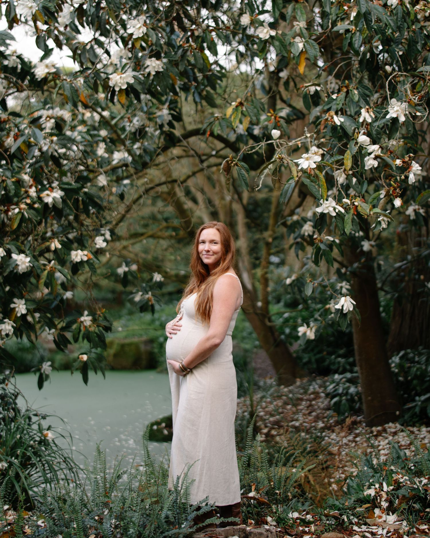 A person in a beige sleeveless top poses among blooming magnolia trees in a garden setting.