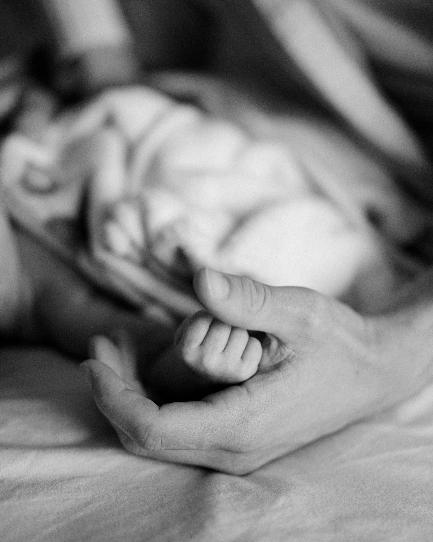 Black and white photo of a newborn baby grasping an adult finger, lying on a soft bed.