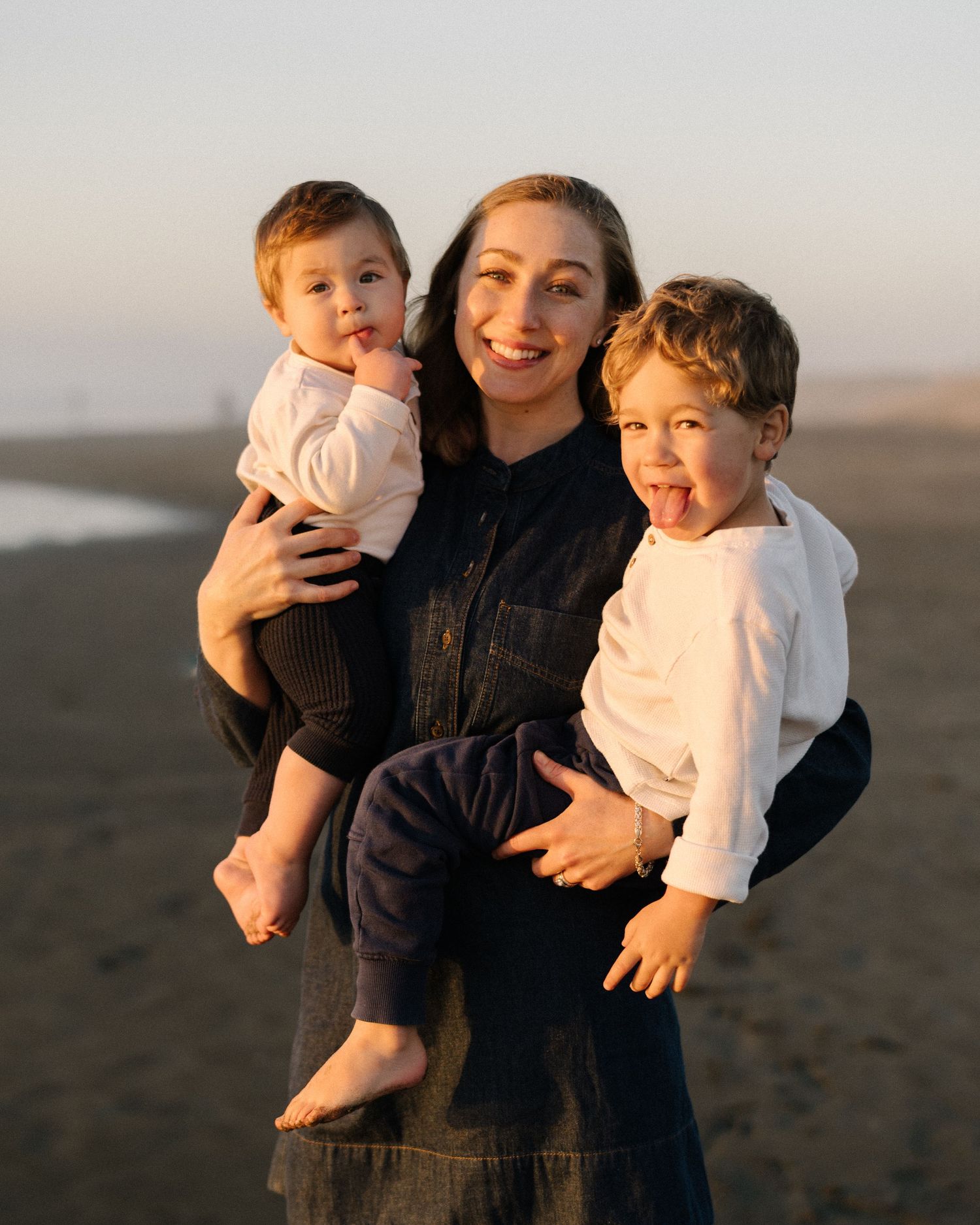 A smiling woman holds two young children on a sunny beach, all dressed casually in a warm golden light.