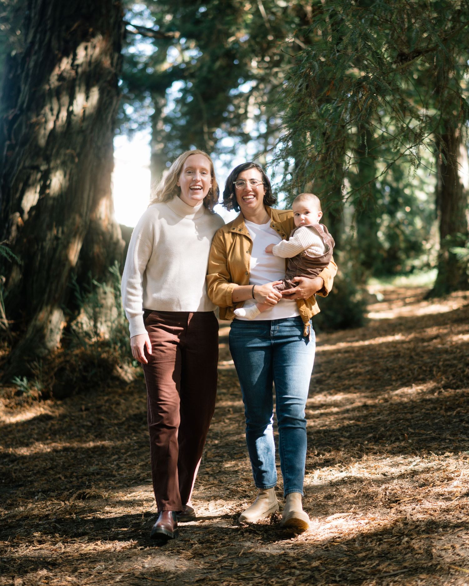 A family enjoys a sunny afternoon walk together on a forest trail surrounded by tall evergreen trees.
