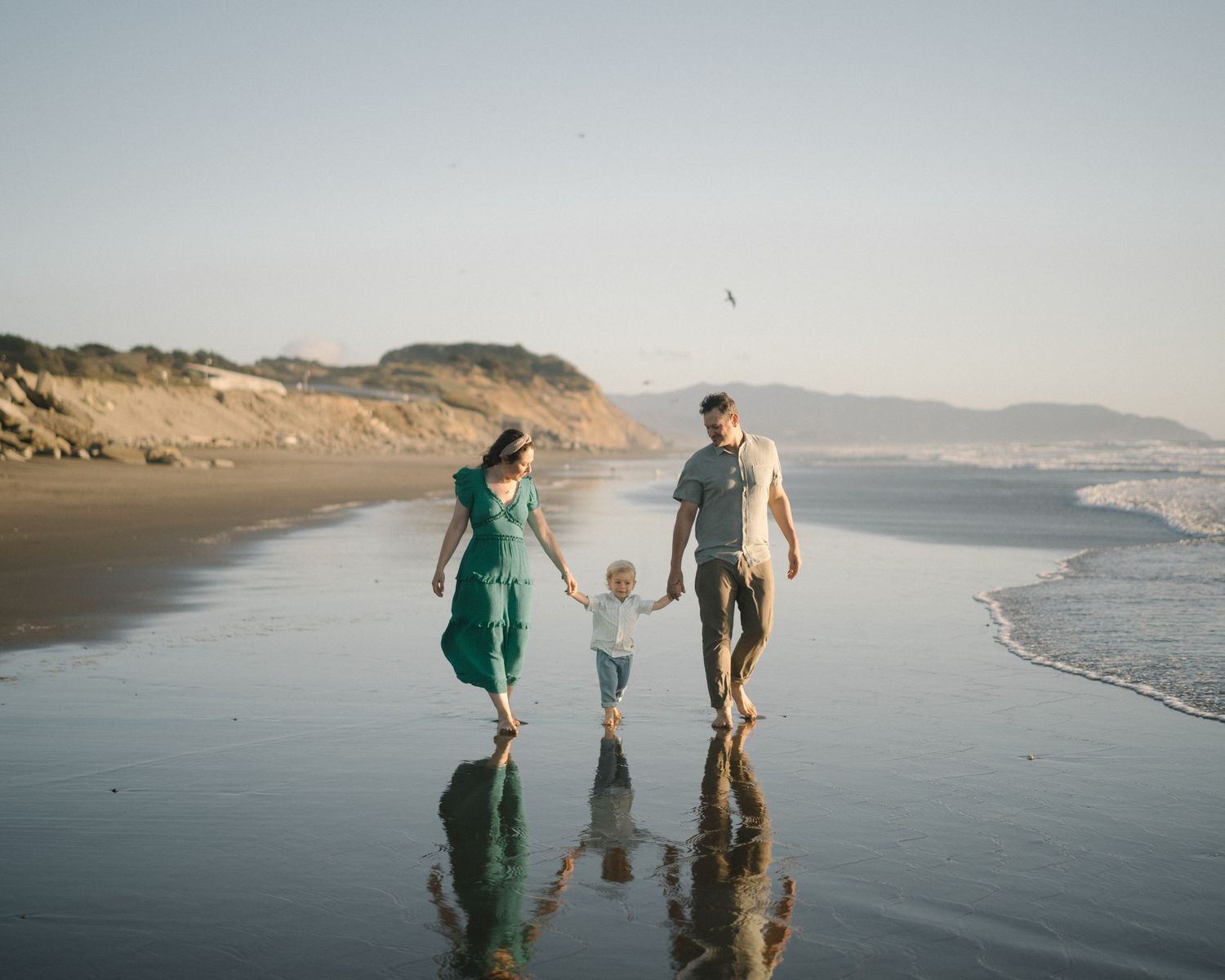 Family strolling on wet sand beach with reflections in water during golden hour.