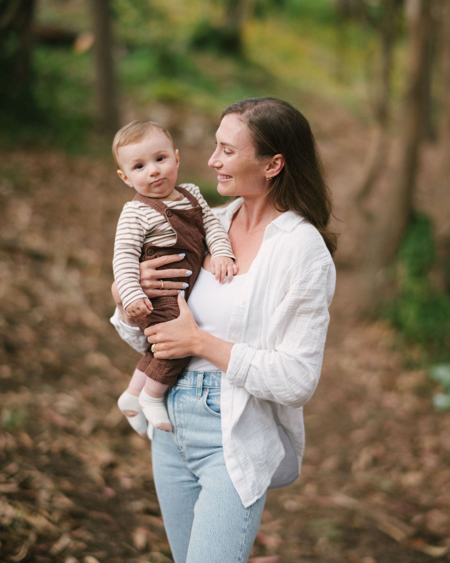 A smiling mother holds her baby in a sunlit forest, both dressed casually in white and brown tones.