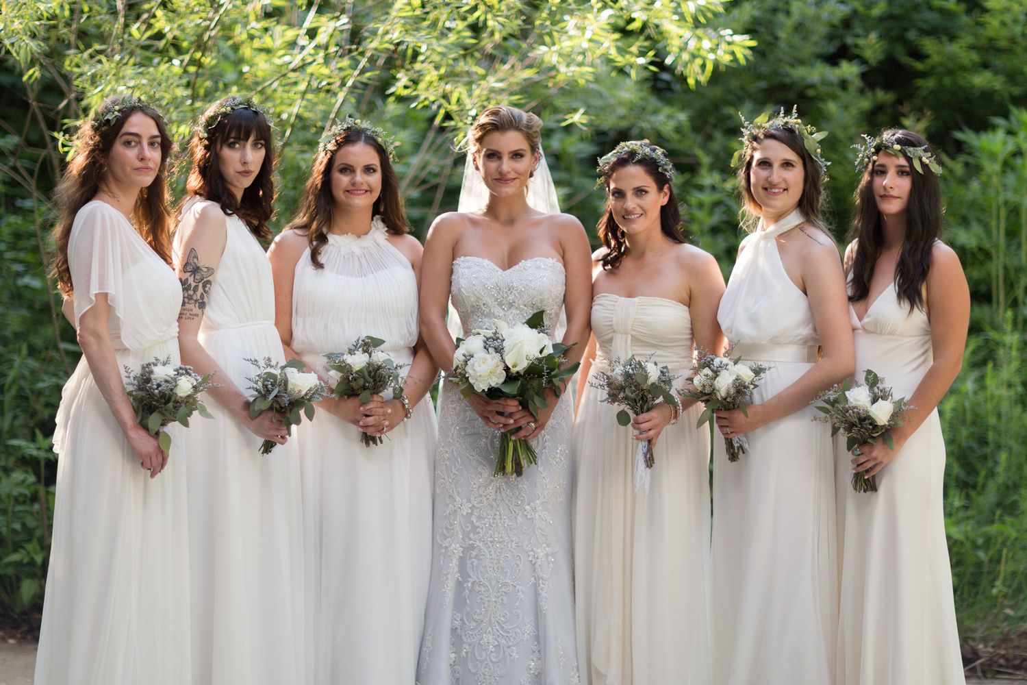 A row of bridesmaids in white dresses holding bouquets stand together in an outdoor garden setting.