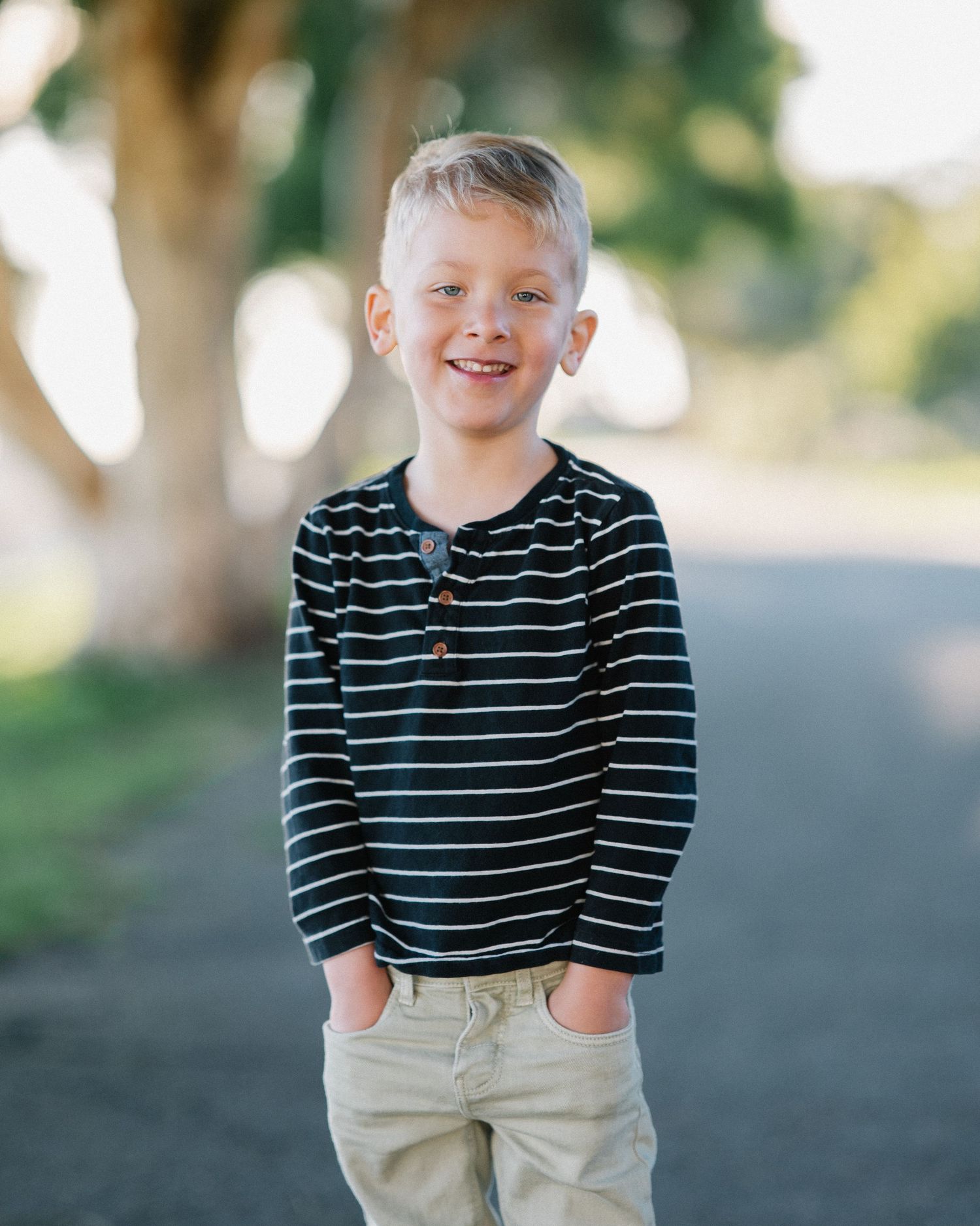 Smiling young boy with blonde hair wearing a striped long-sleeve shirt and khaki pants standing on a path.
