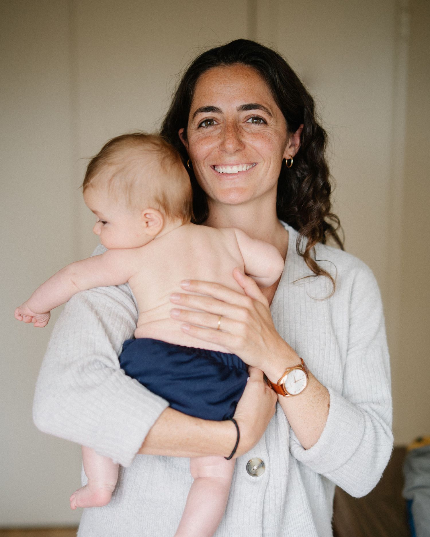 Smiling woman holding a baby in navy blue diaper, wearing a light gray cardigan and rose gold watch.