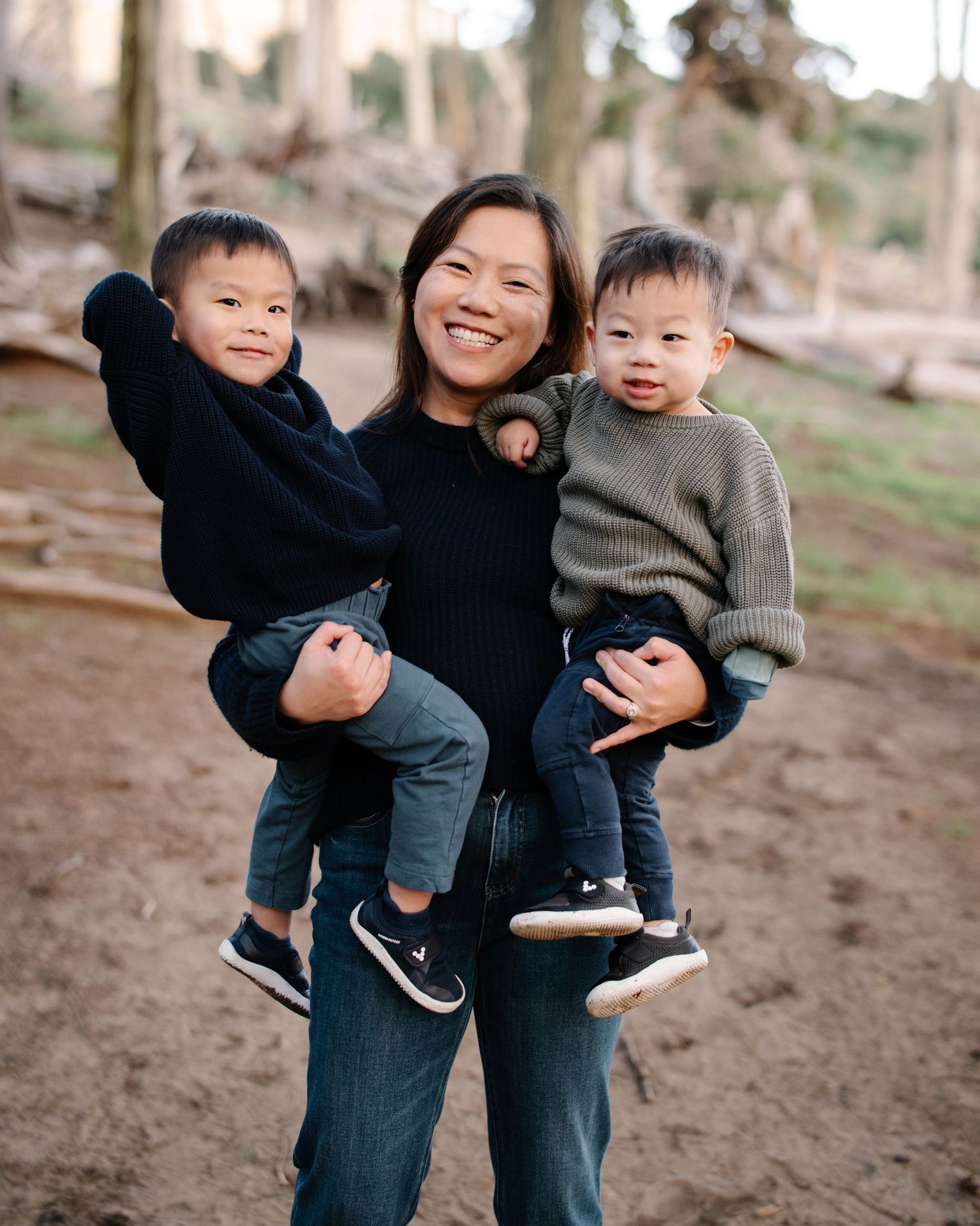Smiling woman holding two young toddler boys outdoors in a wooded park setting.