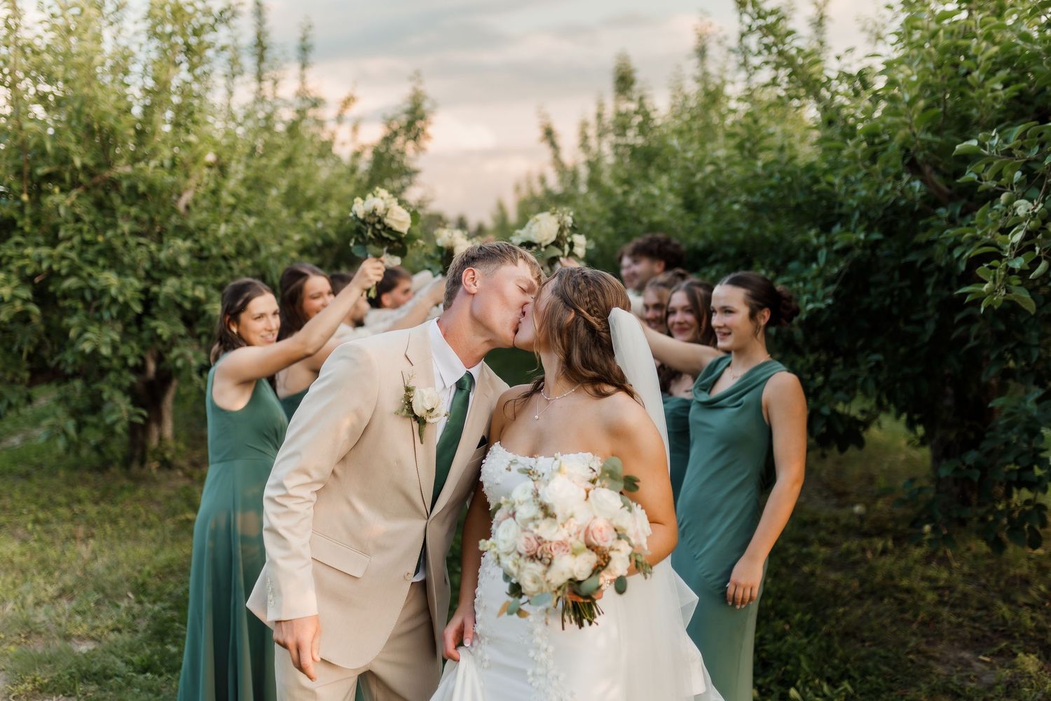 Bride and groom kissing surrounded by wedding party in sage green dresses and tan suits in an orchard.