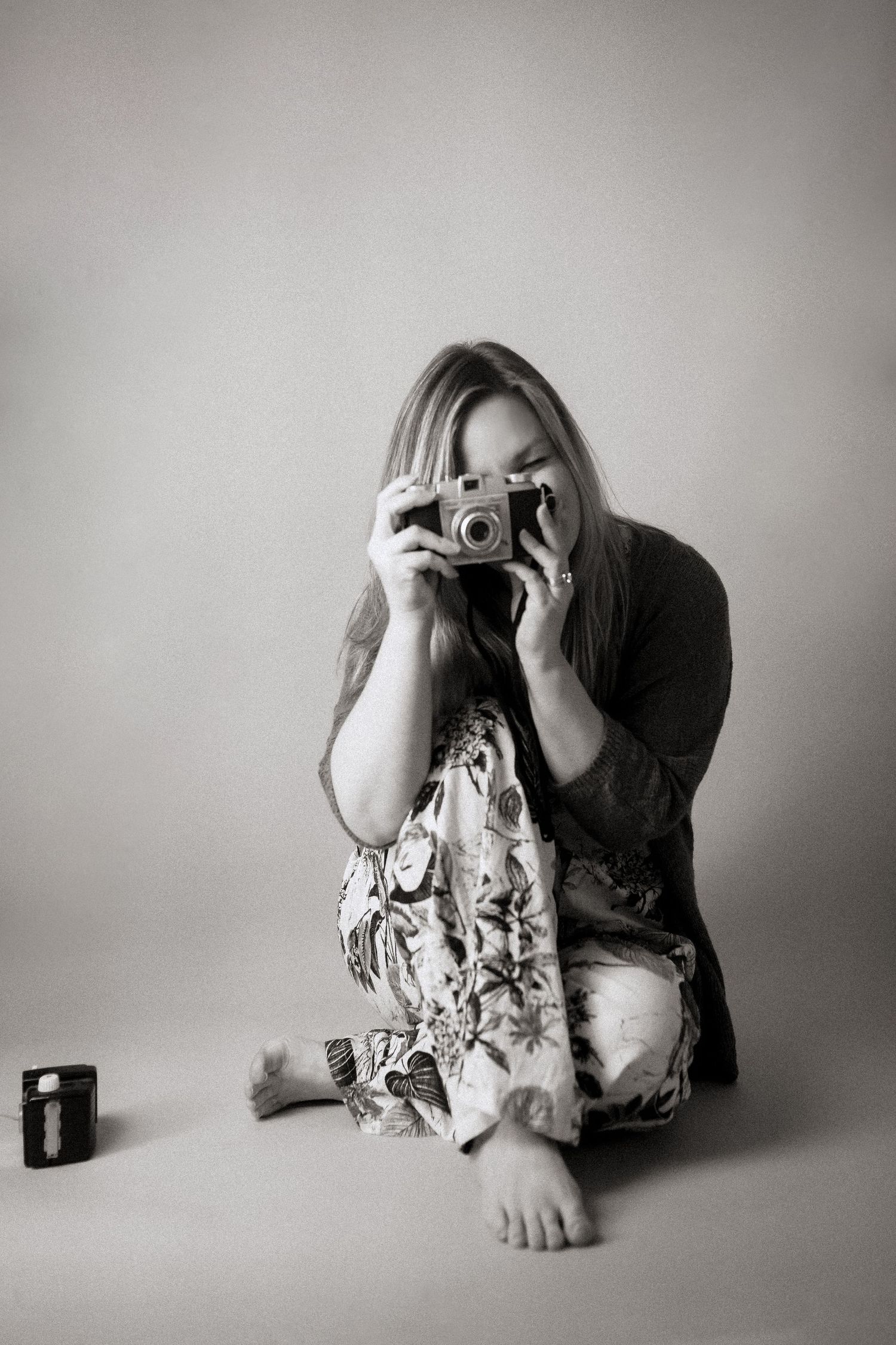A photographer in floral dress sits cross-legged on the floor taking a photo with a vintage film camera.