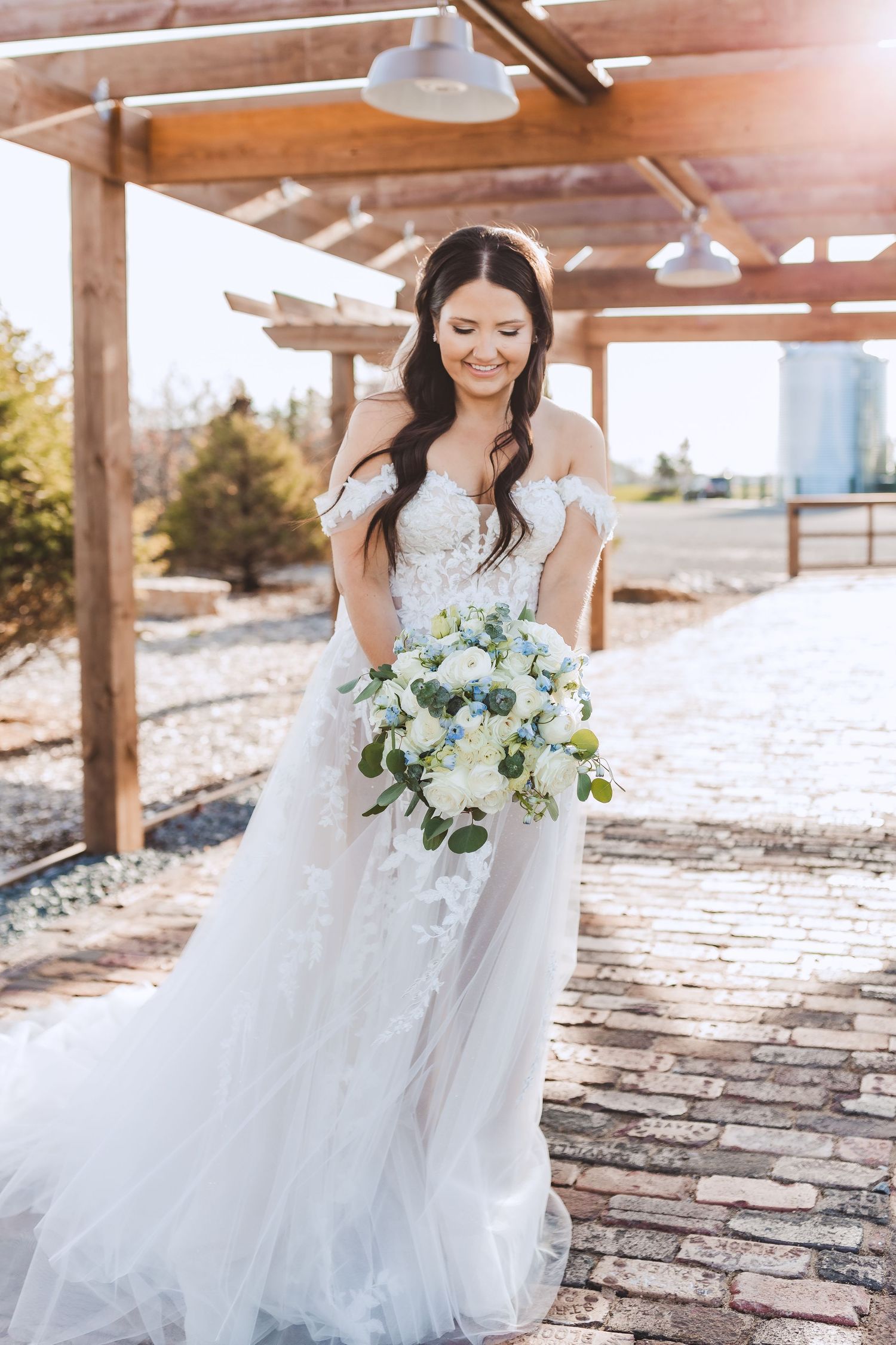 A smiling bride in an off-shoulder white tulle gown holds a blue and white floral bouquet under a wooden pergola.