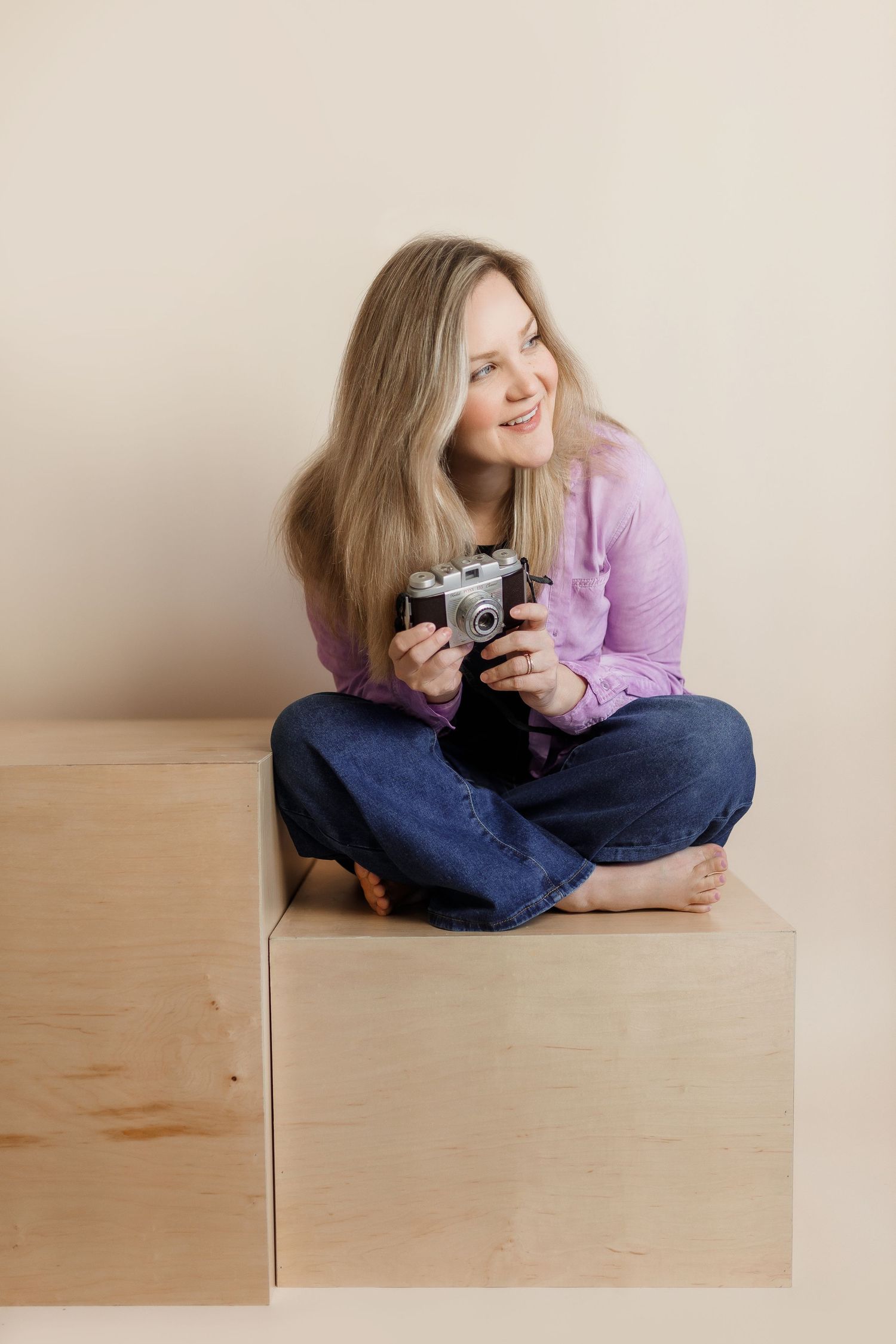 A photographer in a purple sweater and jeans sits cross-legged on a wooden box while holding a vintage camera.
