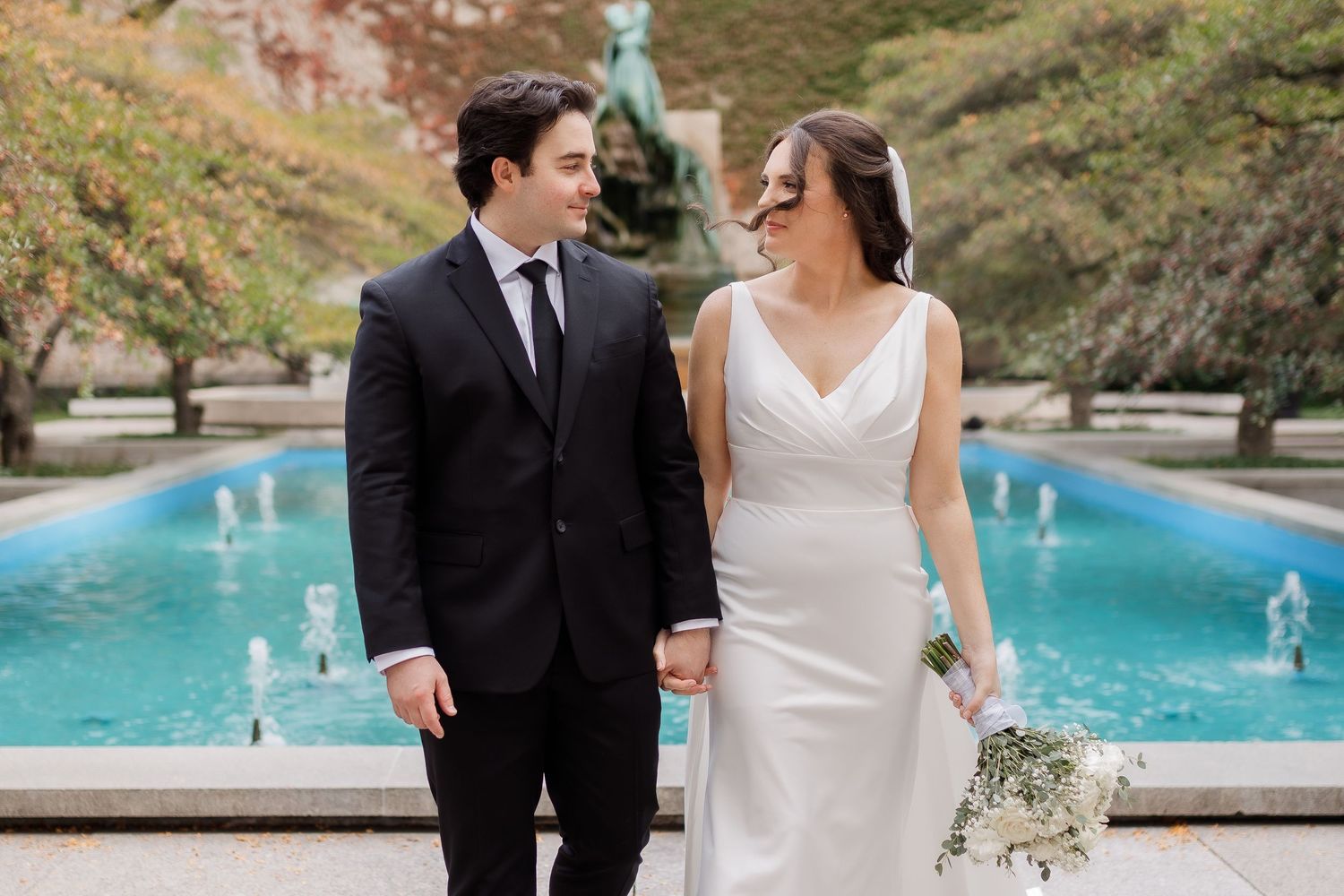 A bride and groom share romantic moments in a garden setting with fountains and lush greenery in the background.