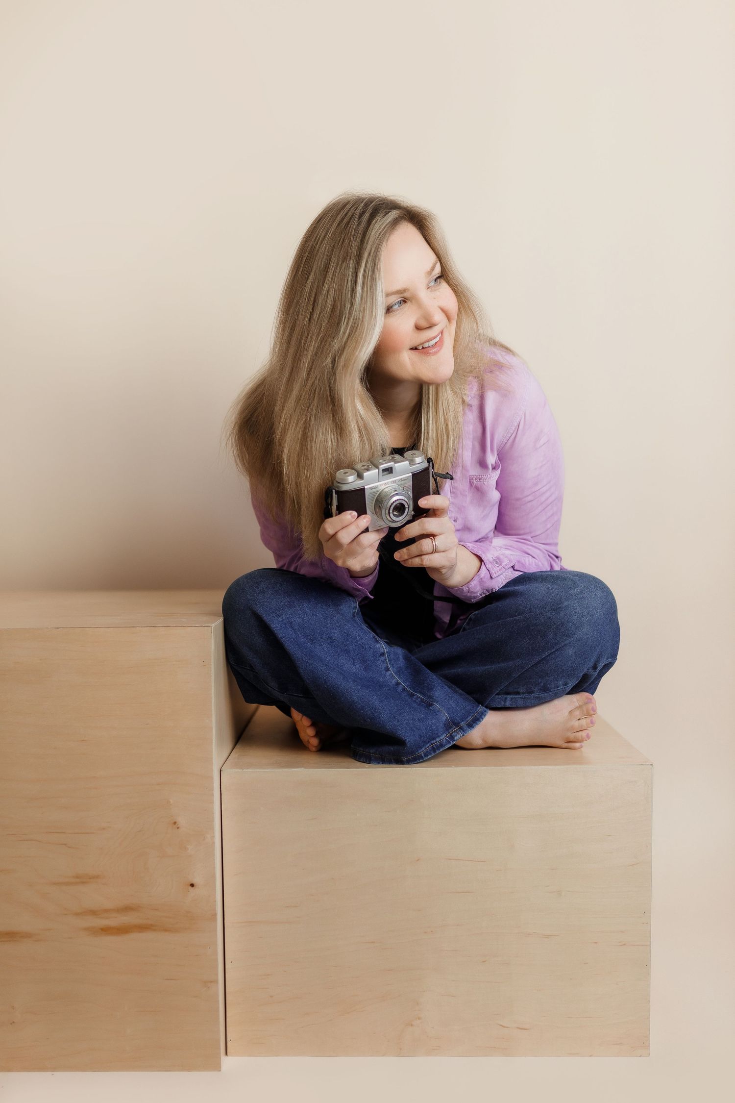 Indiana wedding photographer in a lavender  top and jeans sits cross-legged on a wooden platform while holding a vintage camera.