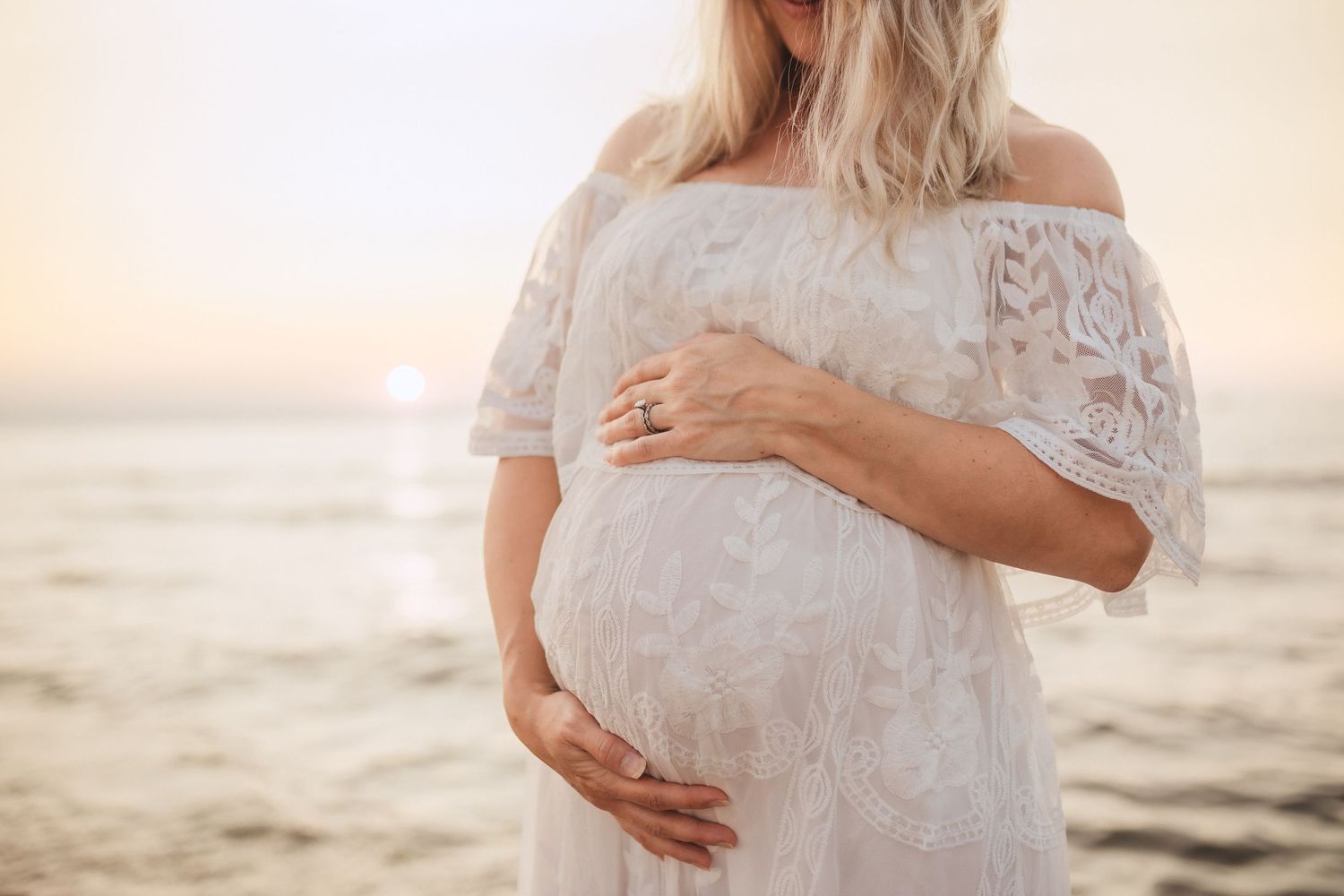 A photographer captures a serene beach sunset moment as hands cradle a pregnant belly in Southwest Michigan at the dunes on Lake Michigan