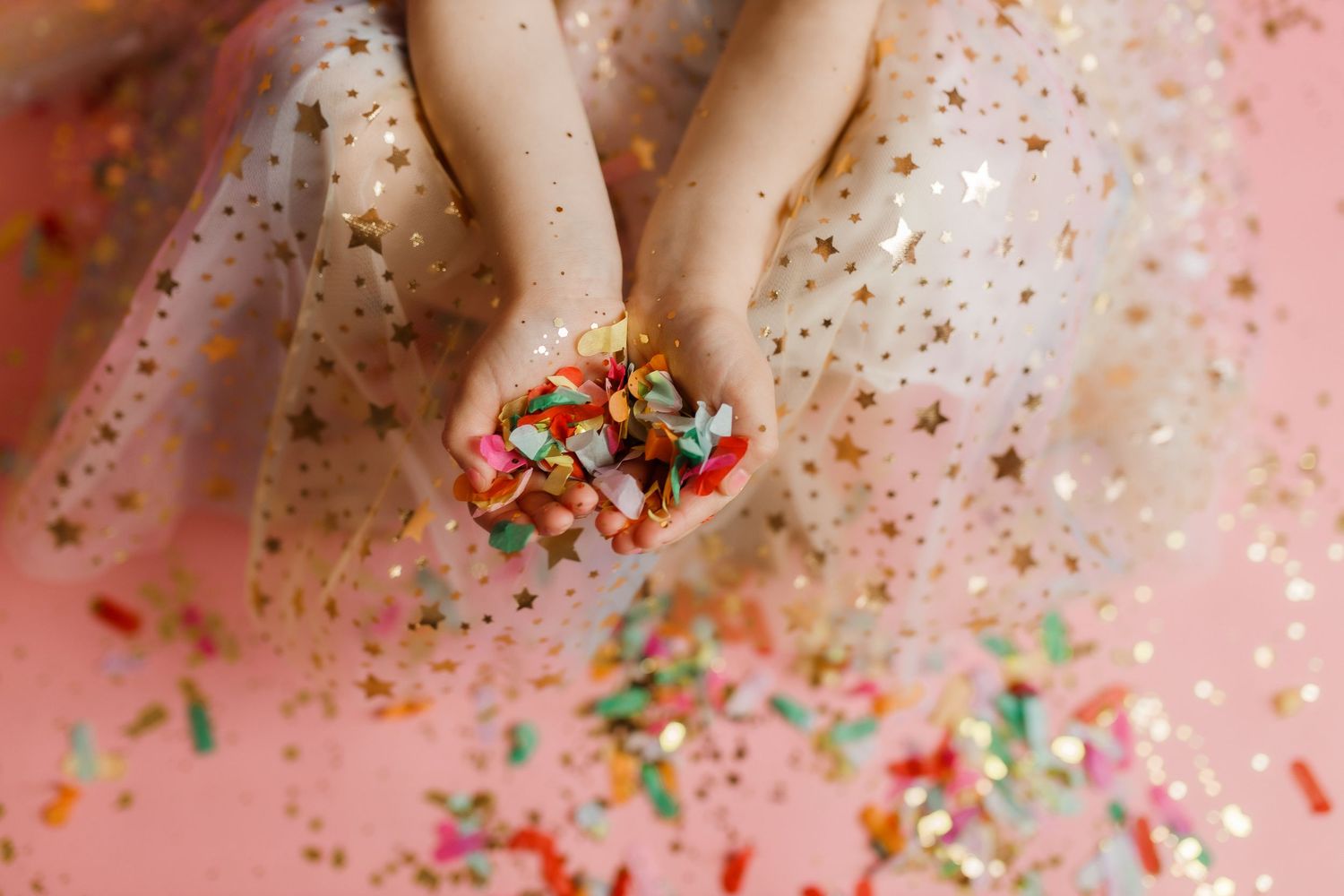 Hands sprinkle colorful confetti and glitter over a pink background in a celebratory gesture.
