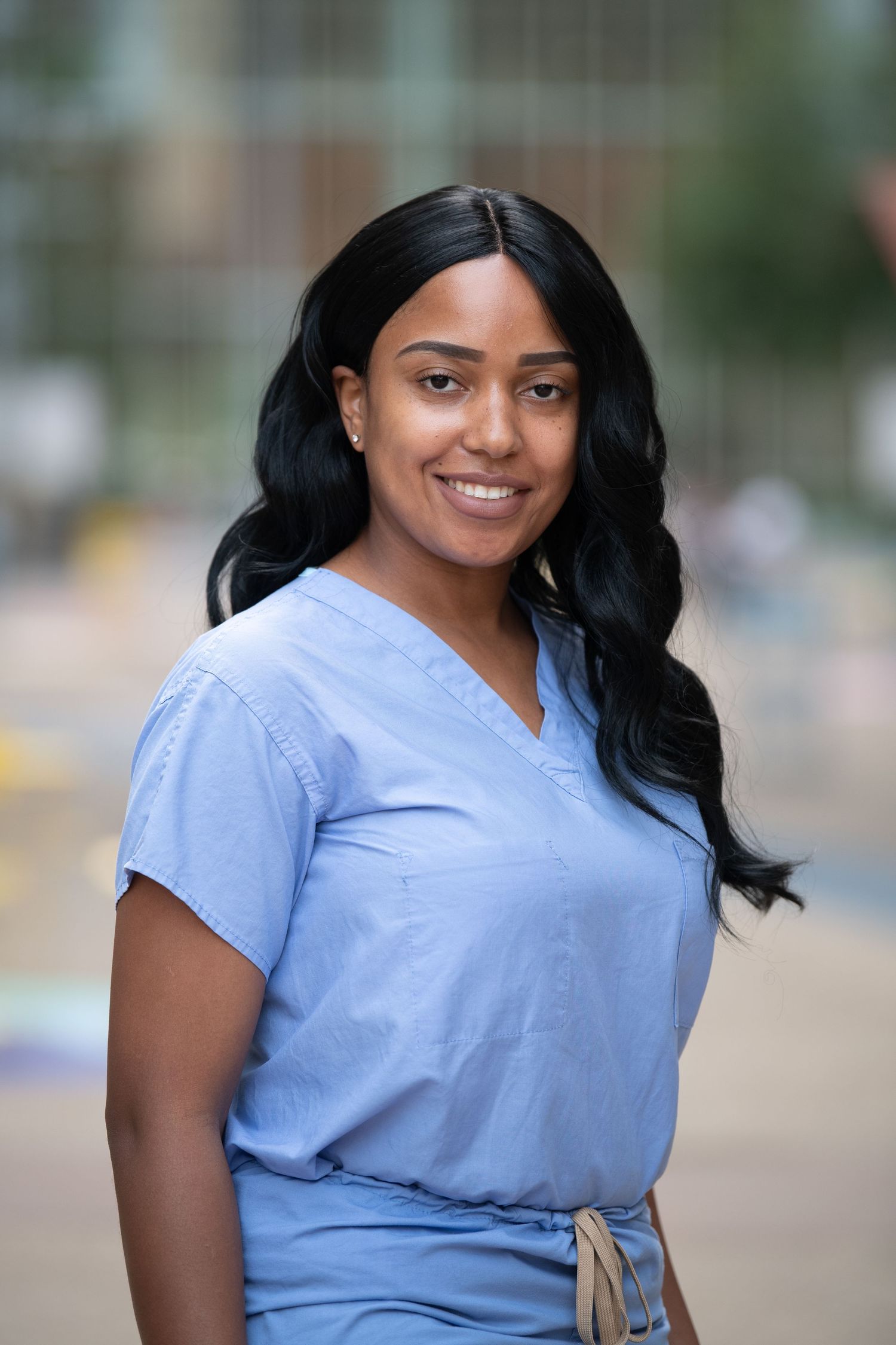 Healthcare professional in light blue scrubs smiling outdoors with soft blurred background.