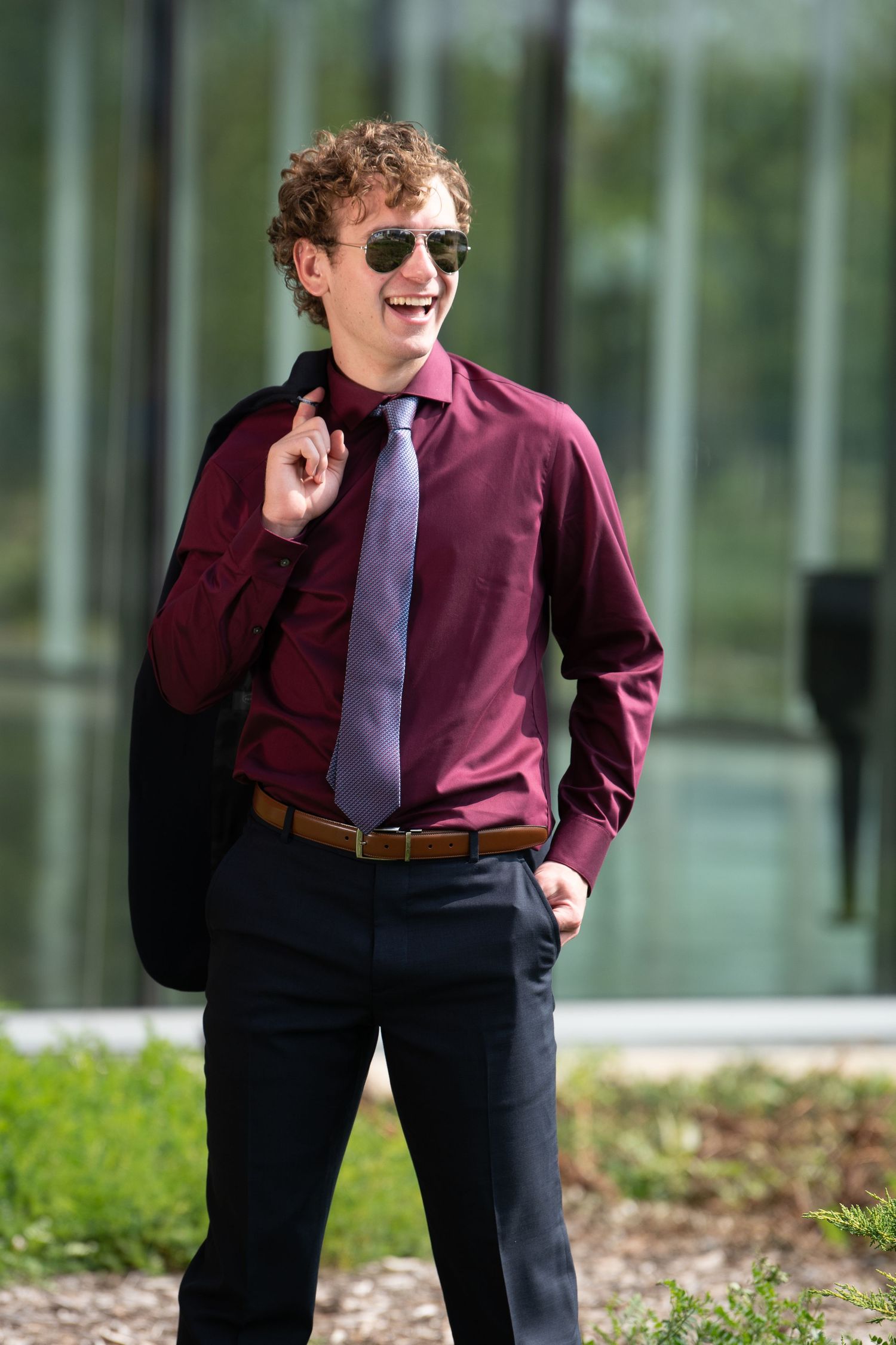 A young professional wearing sunglasses, burgundy dress shirt and purple tie smiles while standing outdoors near a building.