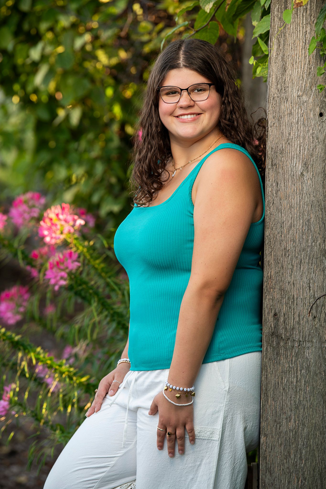 Outdoor portrait with teal tank top and white pants standing by tree with pink flowers blooming in background.