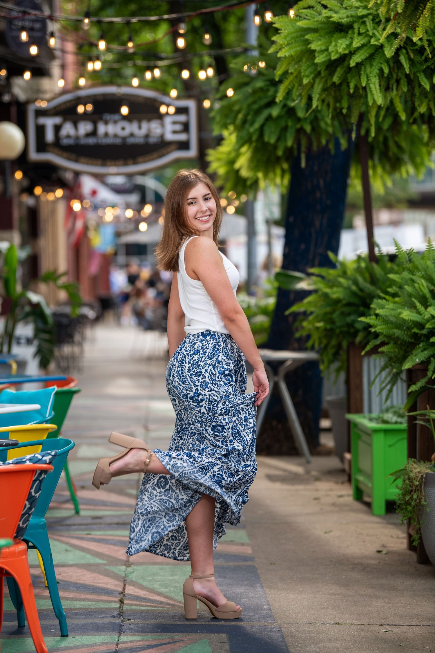 Woman in white top and patterned maxi skirt poses by outdoor restaurant with string lights and hanging plants.