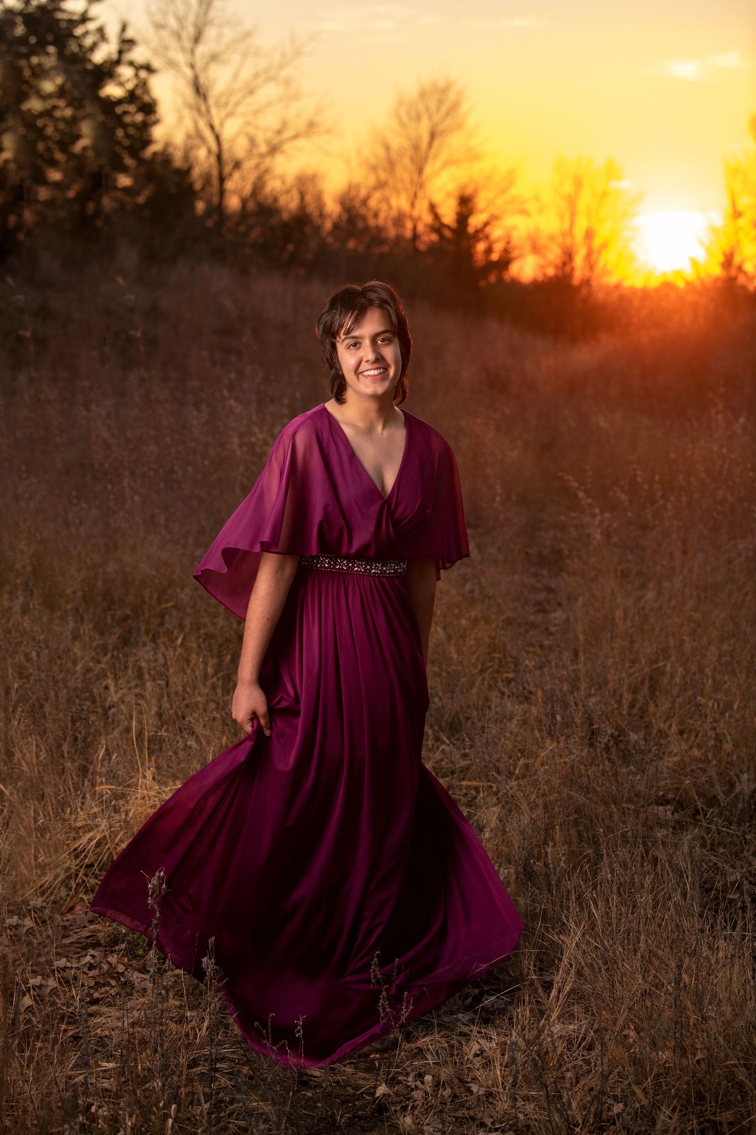 A person in a flowing burgundy dress stands in a field at sunset with golden light illuminating the background.