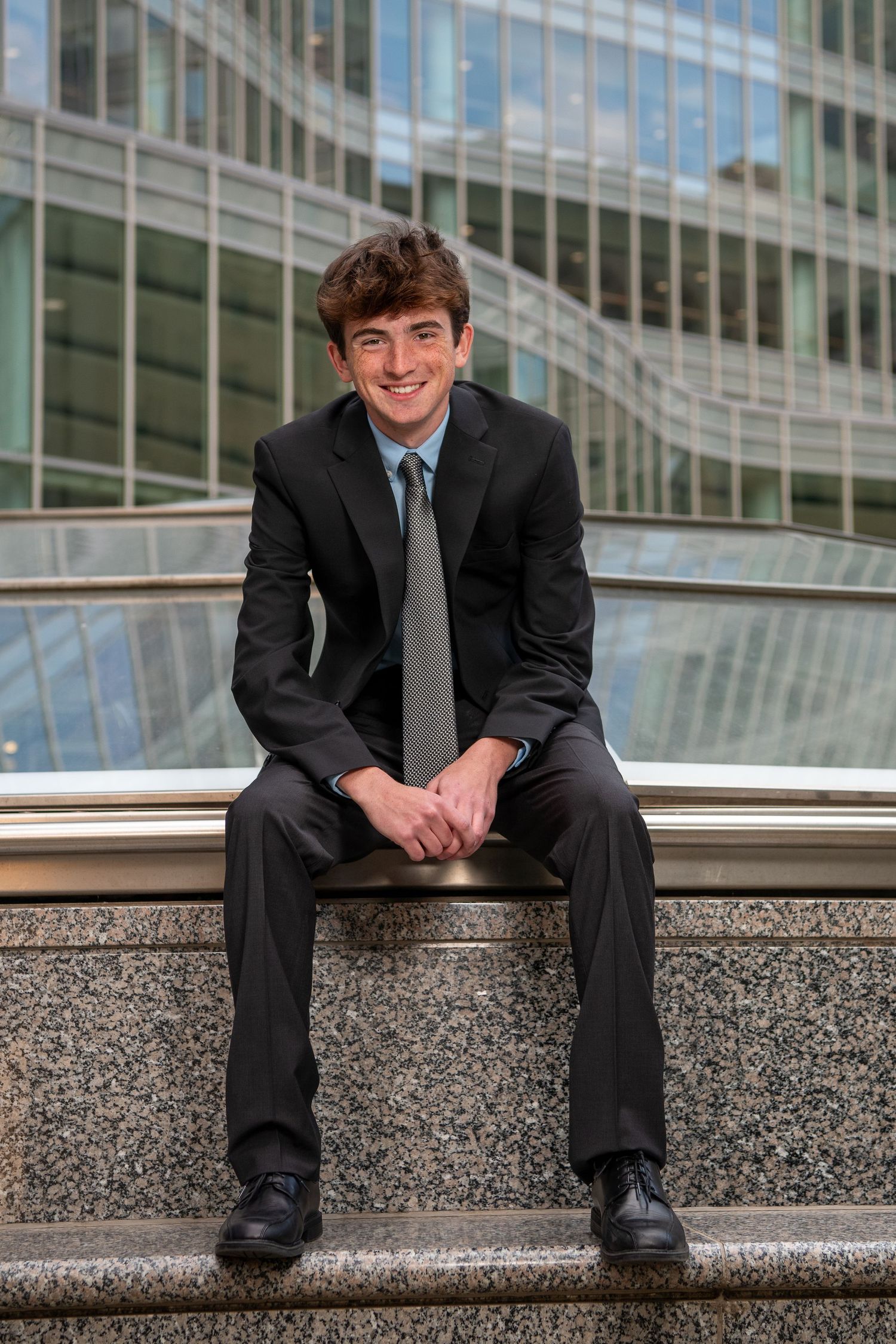 A young professional in a dark suit sits casually on stone steps outside a modern office building with glass windows.