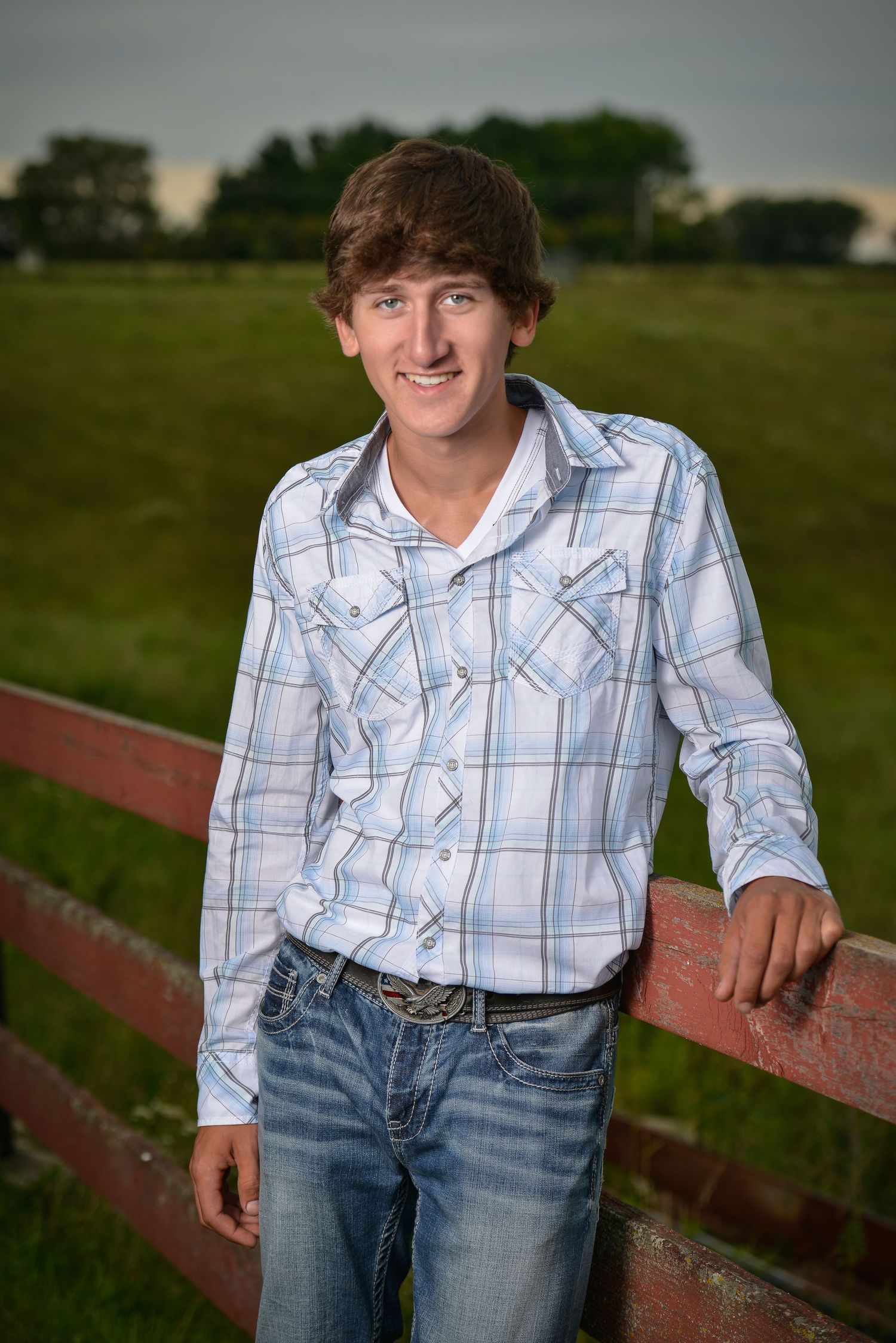 Outdoor senior portrait of someone in a plaid button-down shirt and jeans leaning against a red fence at sunset.