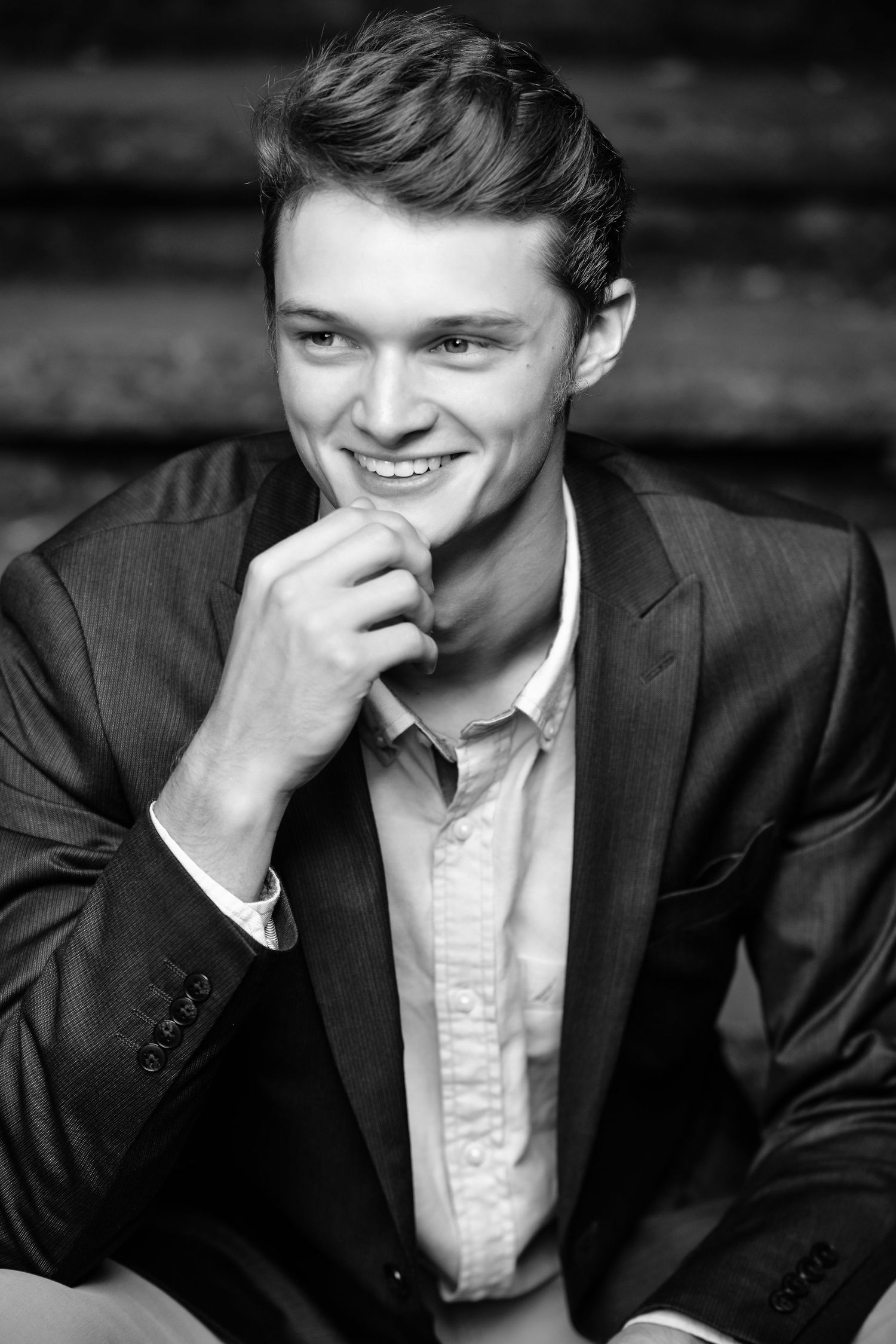 Black and white portrait photo of a high school senior guy in a dark suit jacket smiling warmly with hand on chin.