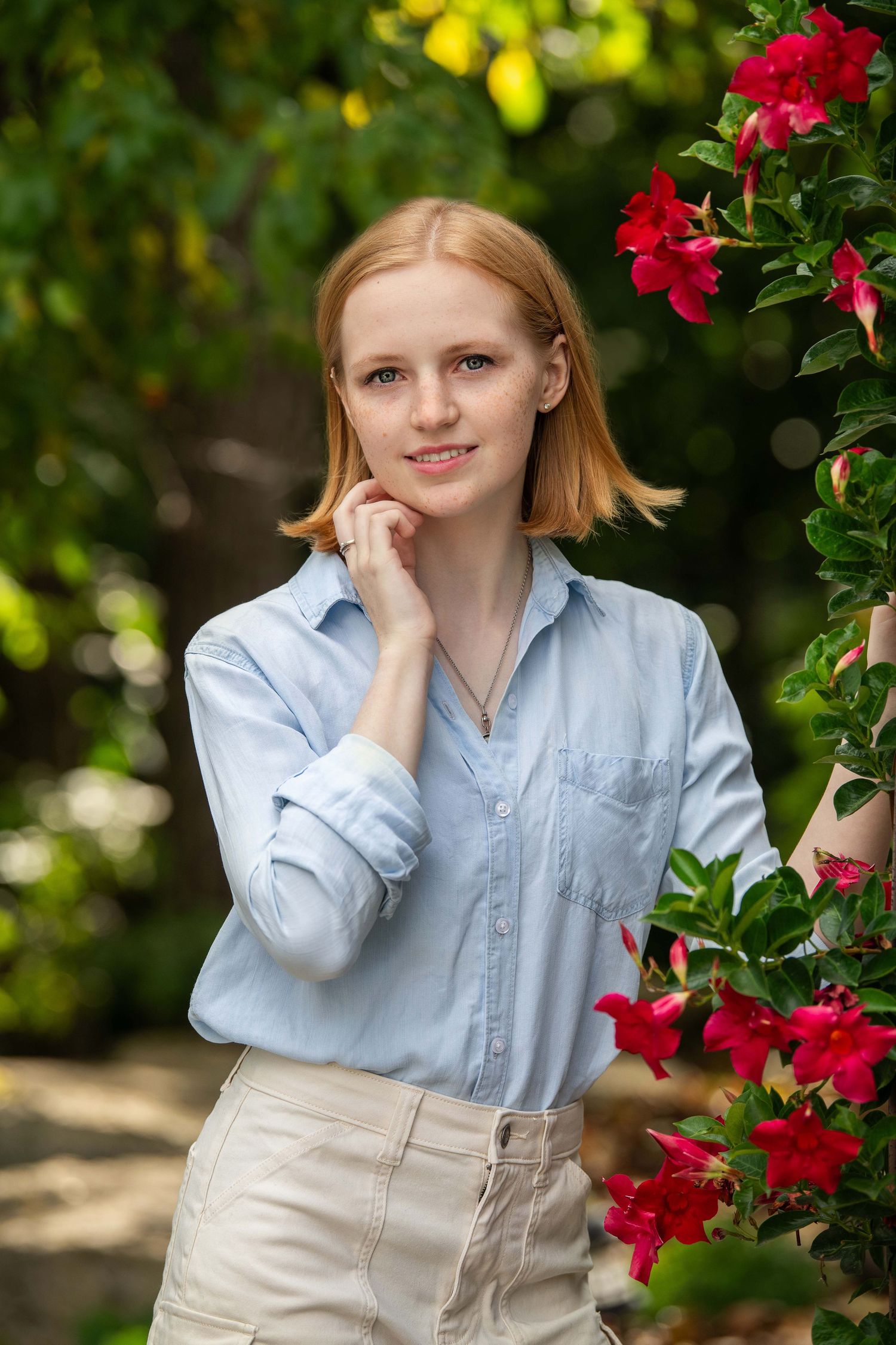 Portrait in a garden setting with vibrant red flowers and natural greenery, wearing a light blue button-down shirt.