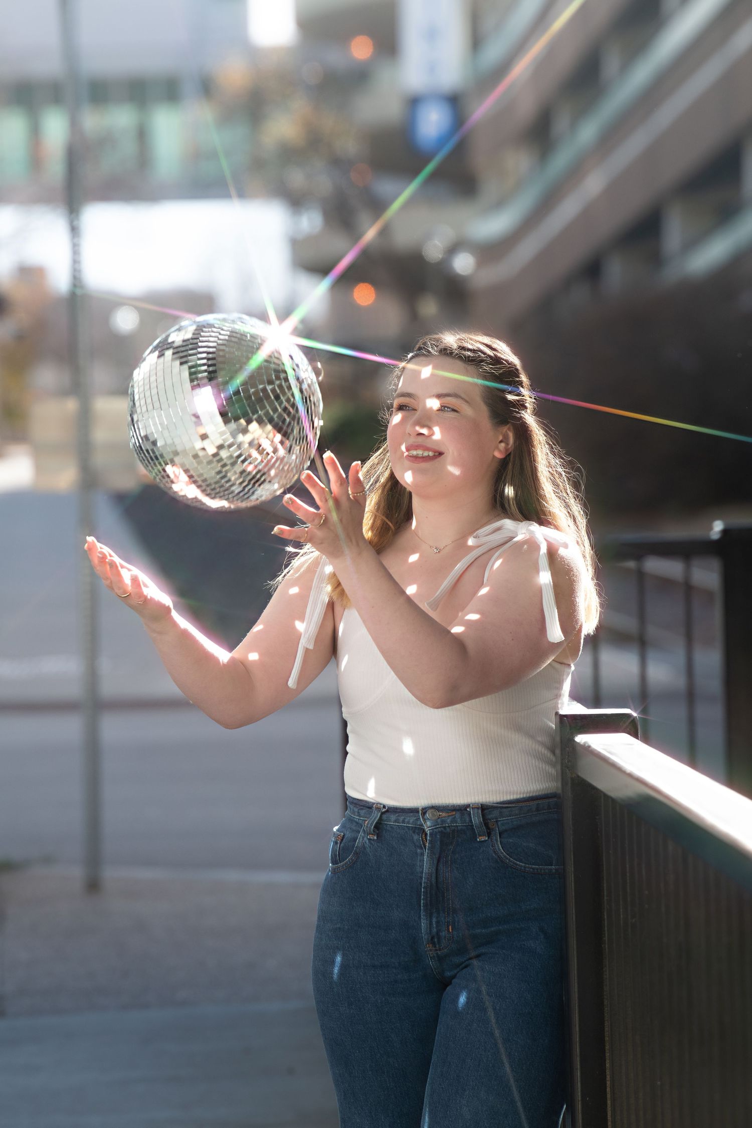 A shimmering disco ball reflects colorful light beams while being held up on an urban balcony or walkway.
