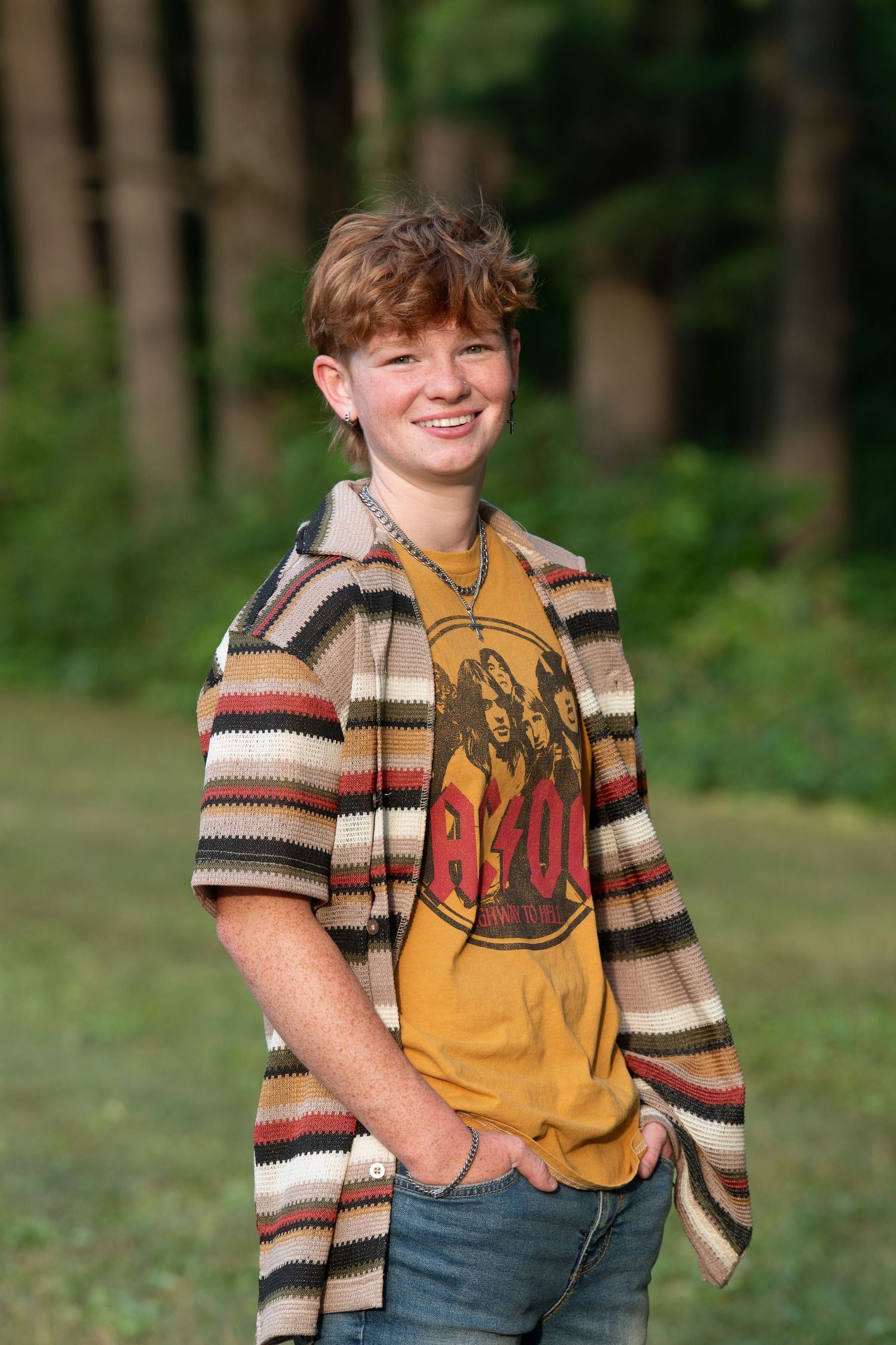 Person wearing a yellow AC/DC t-shirt and striped cardigan poses casually outdoors against a wooded background.