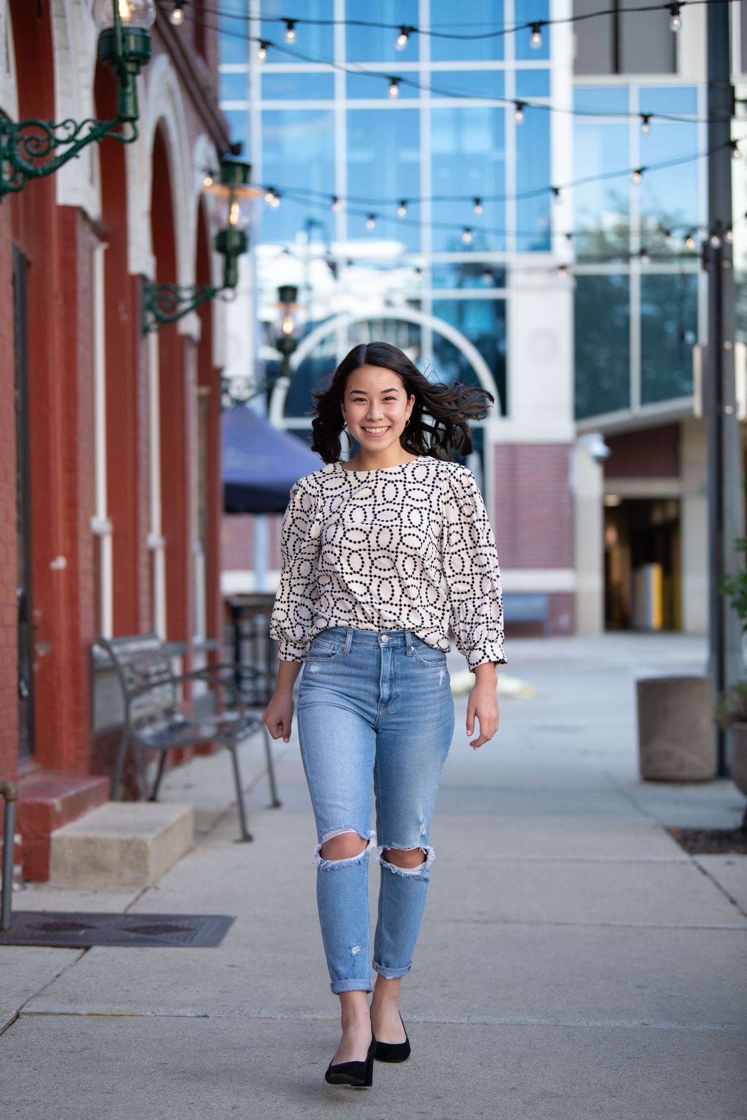 A person in a geometric print top and ripped jeans walks down a street lined with string lights and brick buildings.