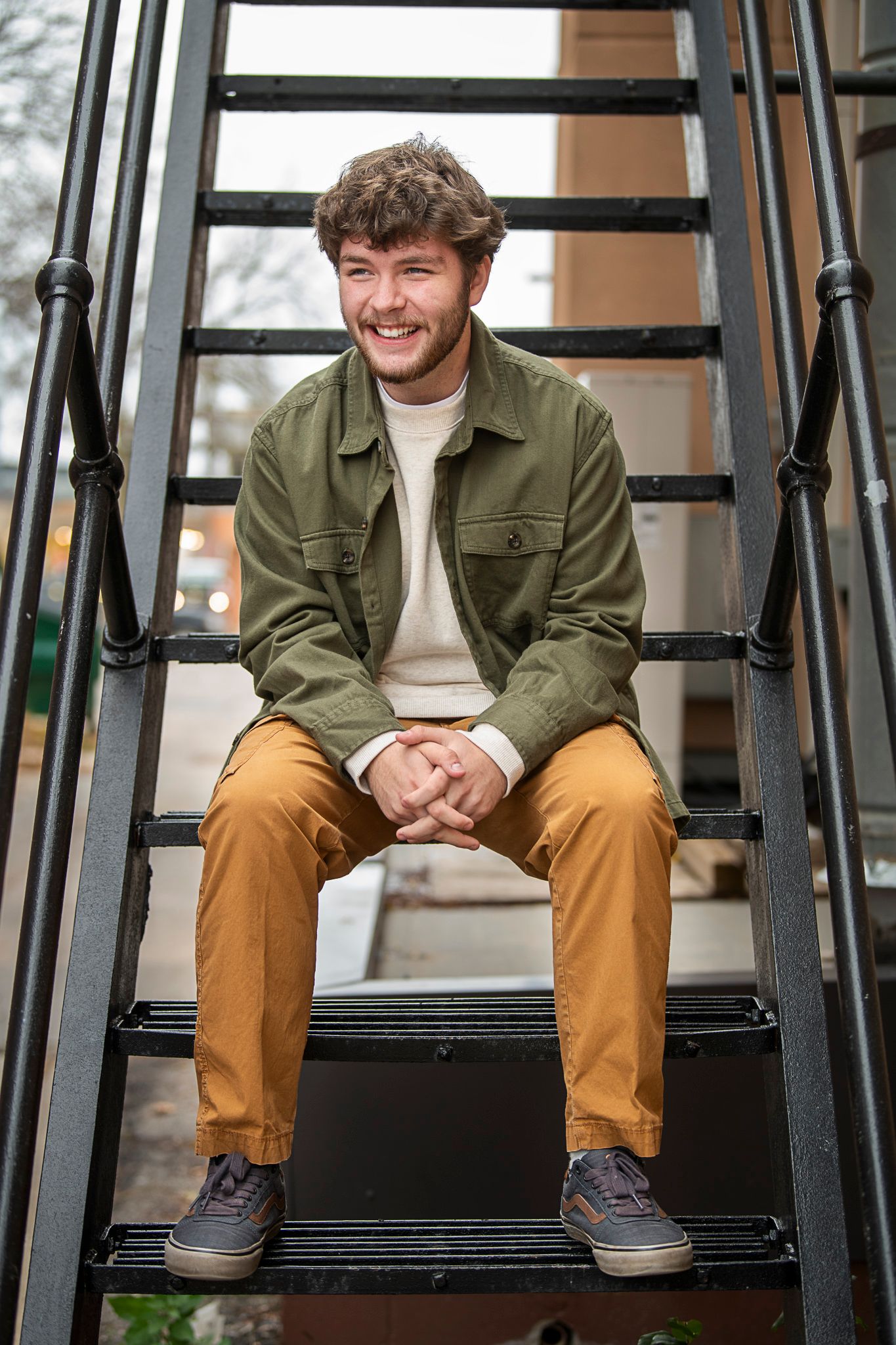 Casually dressed person sitting on black metal stairs wearing olive jacket, beige sweater and brown pants.