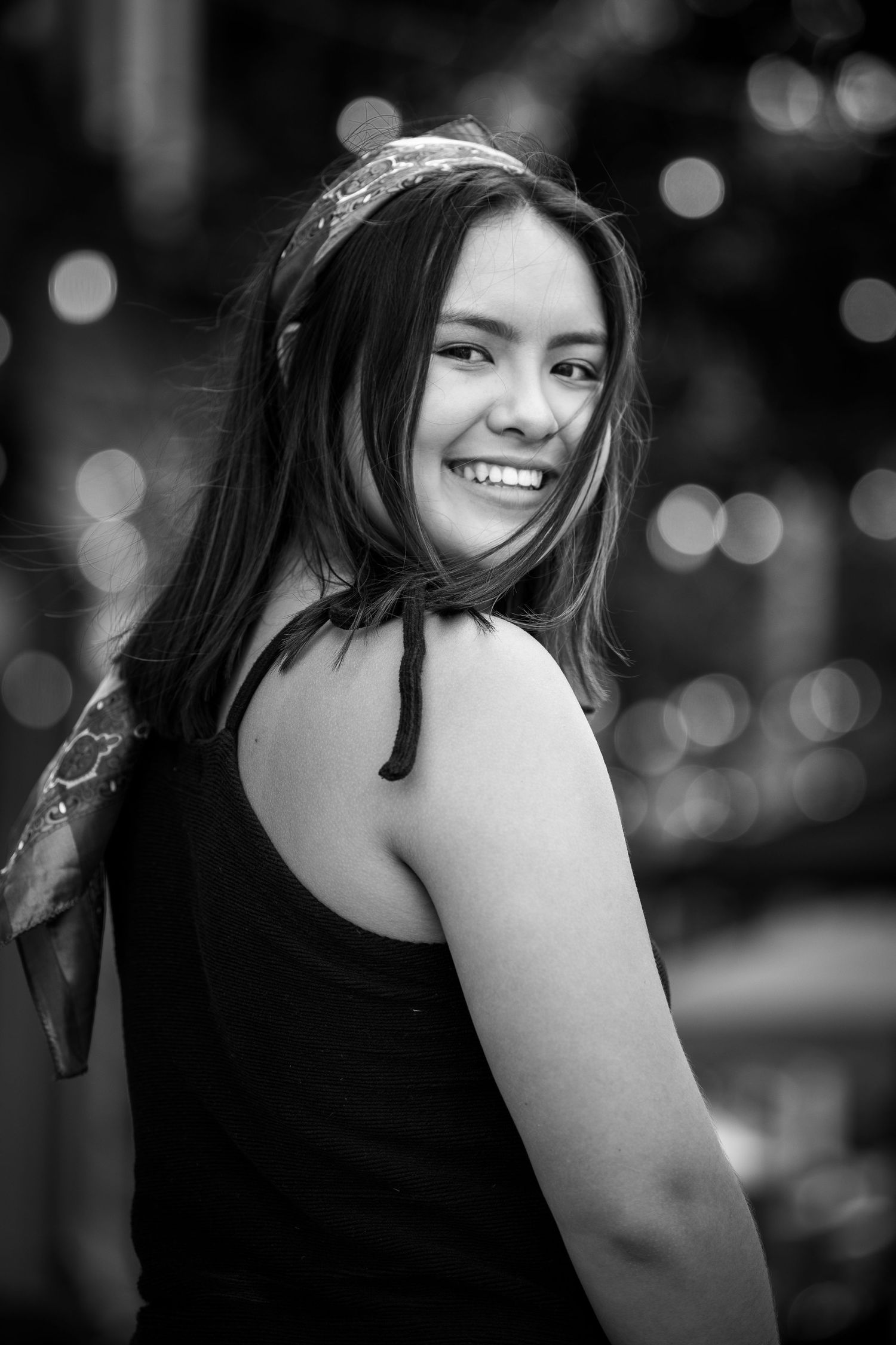 A black and white portrait shows a smiling person in a tank top looking back over their shoulder with bokeh lights in background.