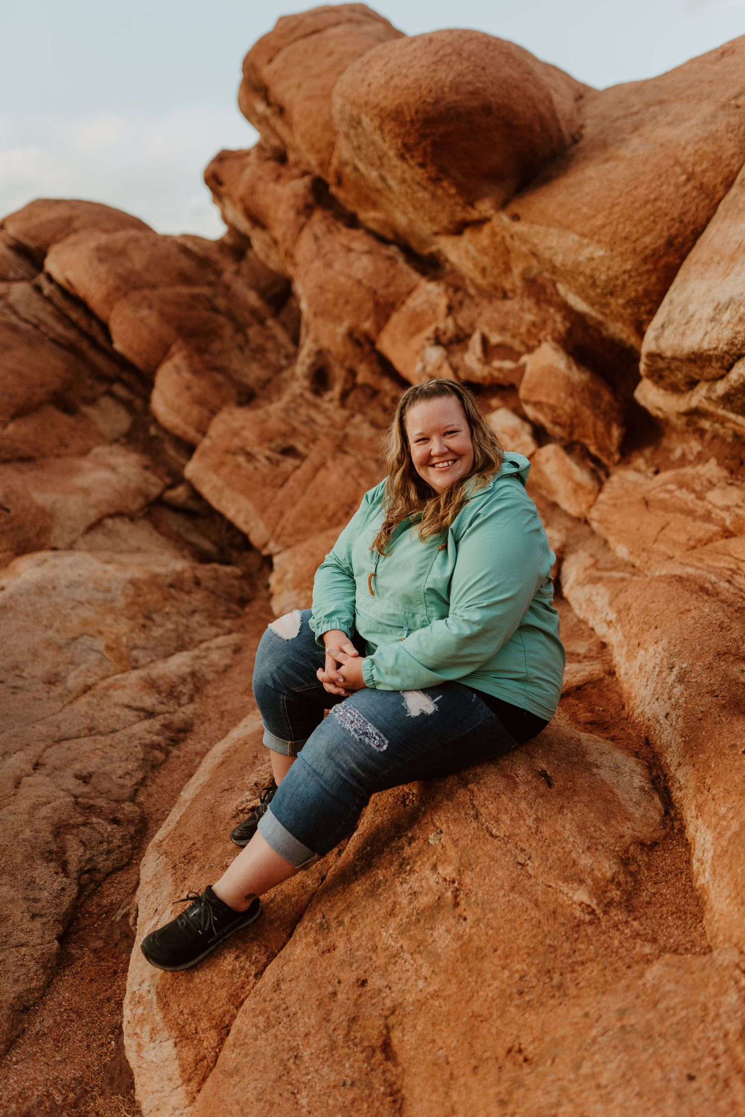 Someone wearing a mint green jacket sits on red sandstone rock formations during sunset.