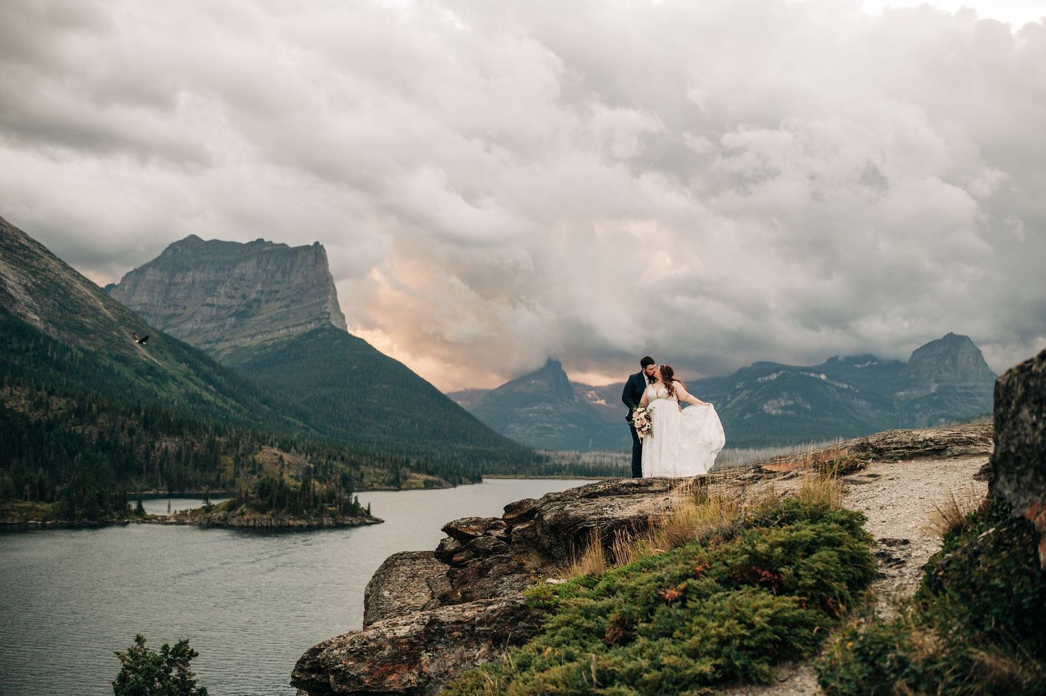 A couple stands on a rocky overlook with a dramatic mountain landscape and lake view at sunset in Glacier National Park.