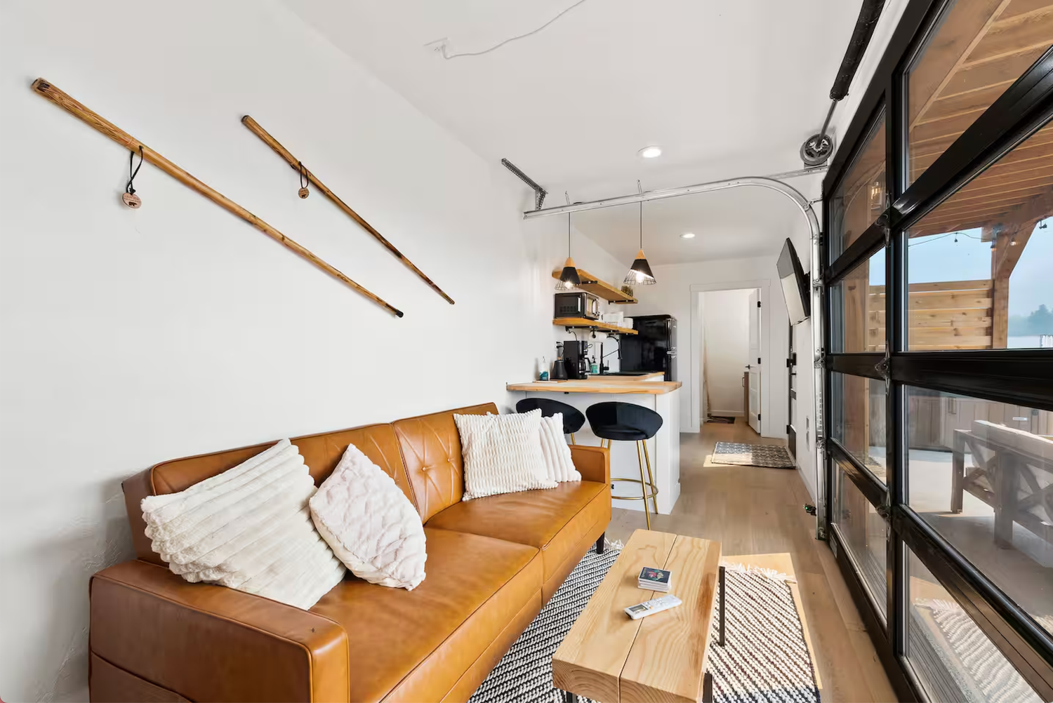 Modern studio apartment interior with tan leather sofa, white pillows, and decorative oars mounted on white wall.