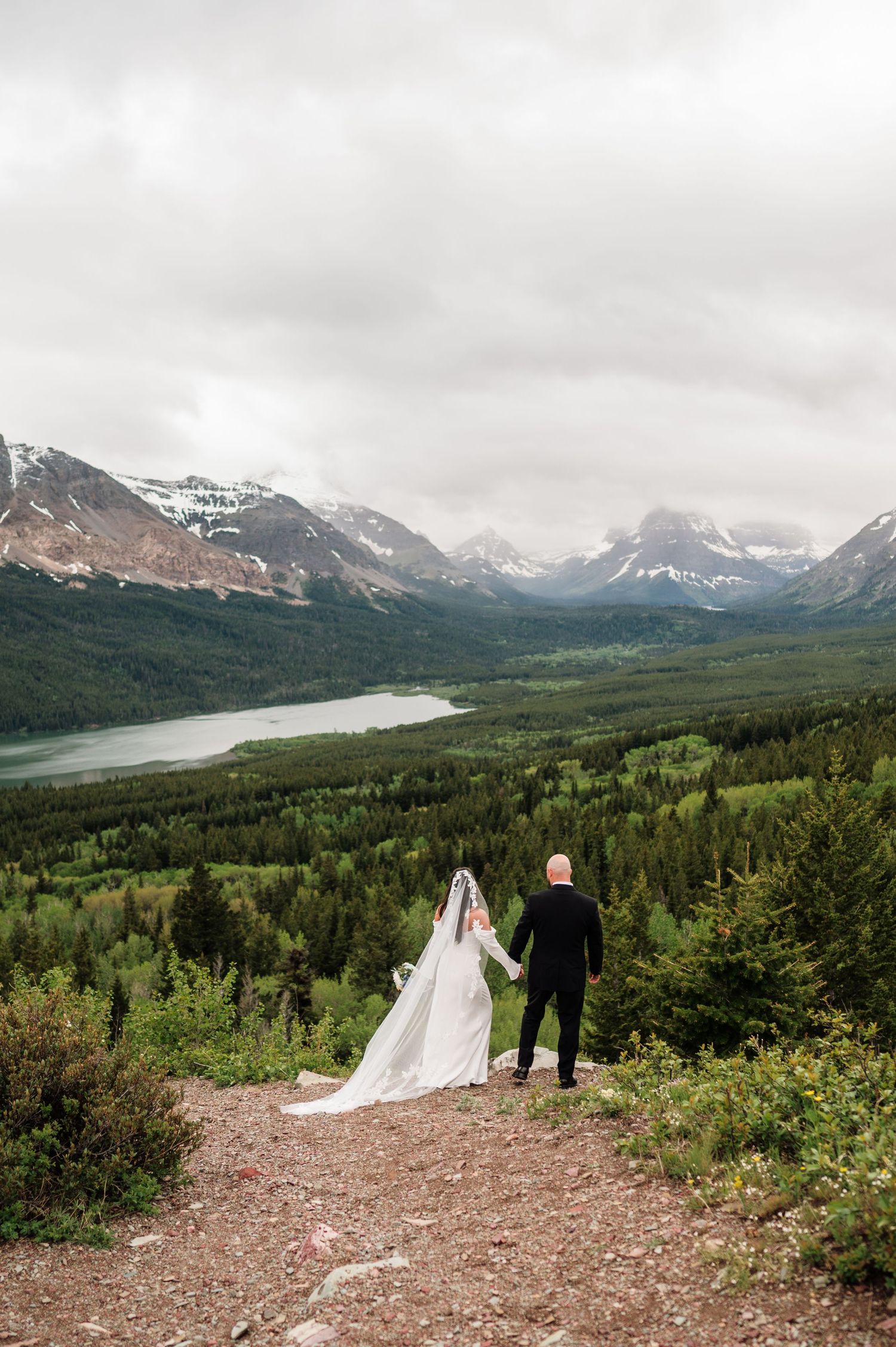 Bride and groom hold hands overlooking a stunning mountain valley with a lake and evergreen forest on their wedding day.