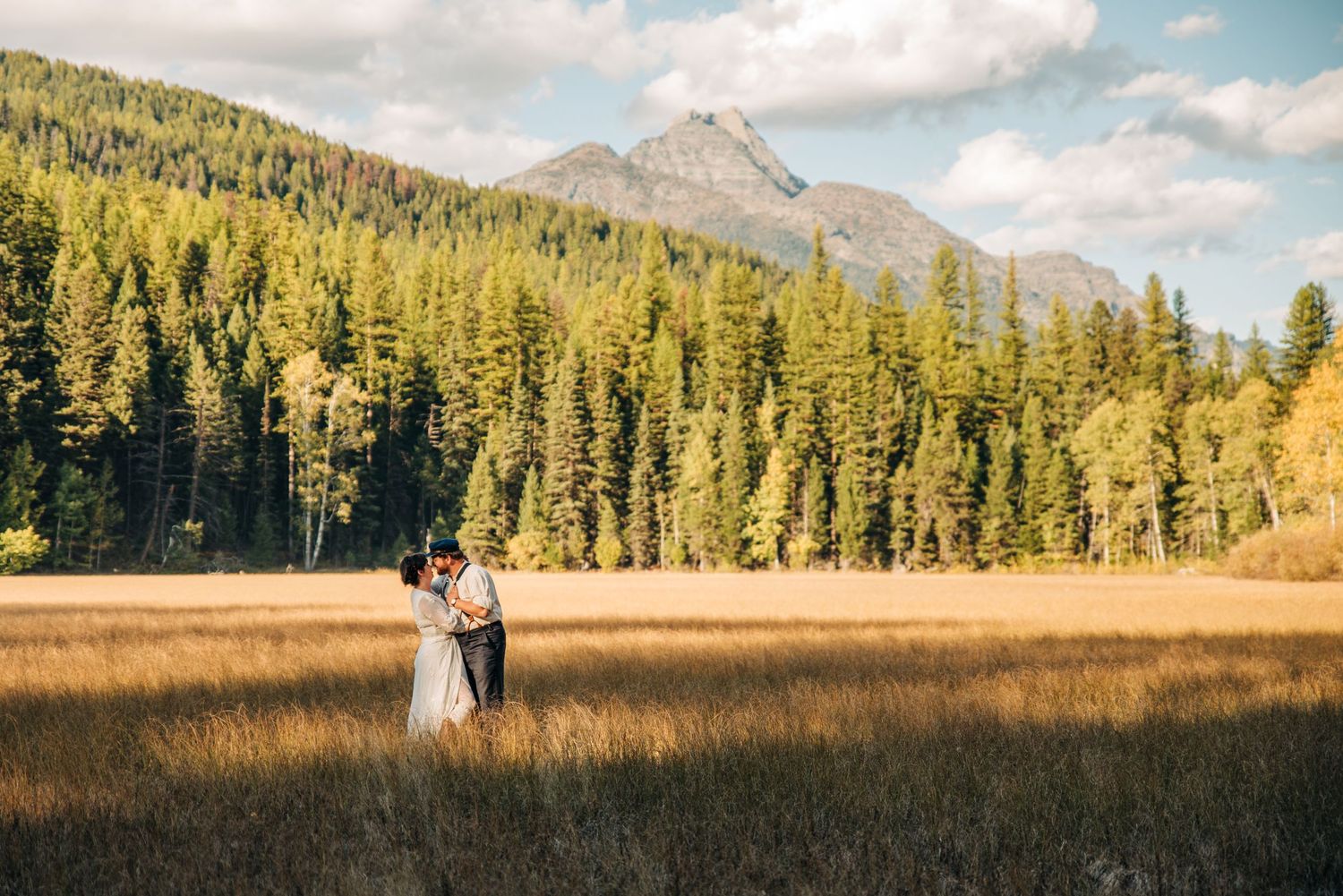 Two figures stand together in a golden meadow surrounded by evergreen forest and mountain peaks in Alberta.