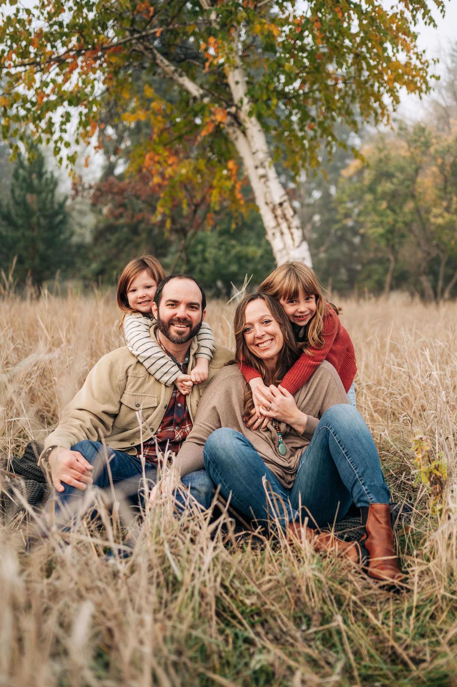 A couple sits together in a tall autumn meadow beneath colorful fall trees on a sunny afternoon.
