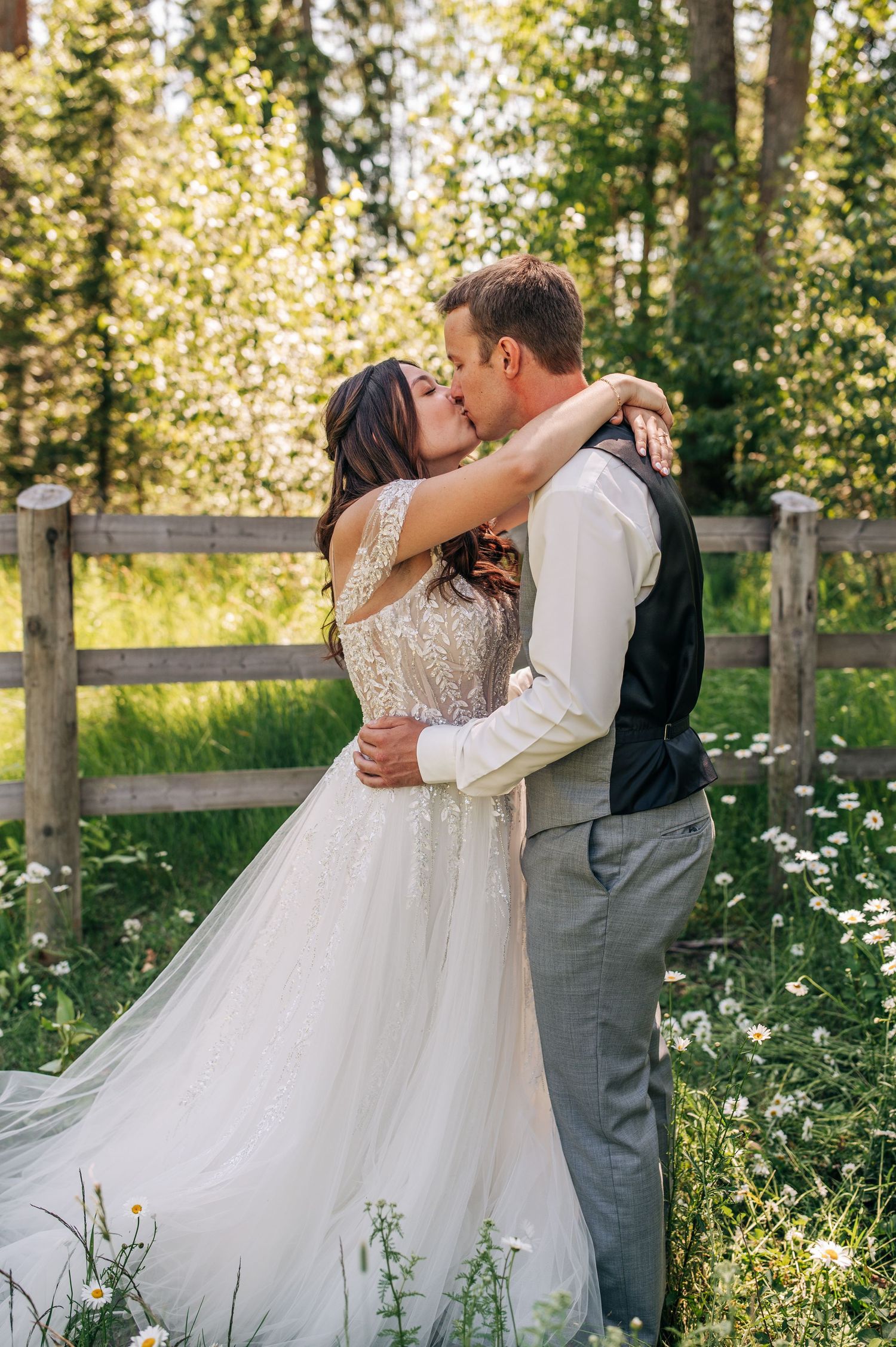 Romantic couple shares kiss in meadow with rustic wooden fence and wildflowers.