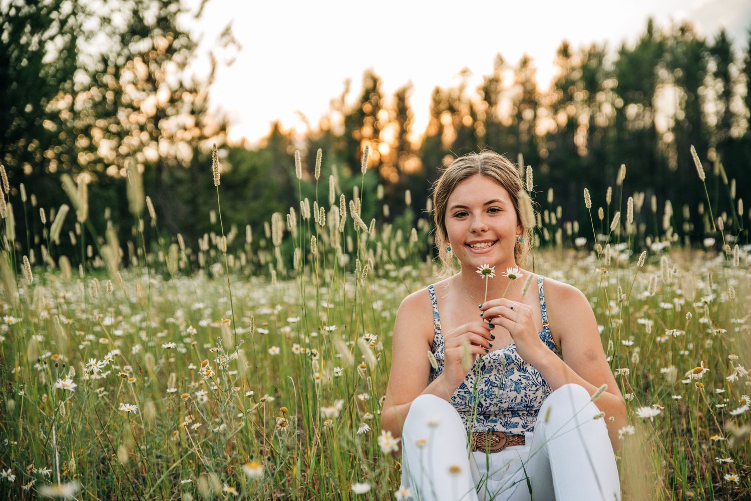 Someone sits in a wildflower meadow at sunset wearing a floral top and white pants while smiling at the camera.
