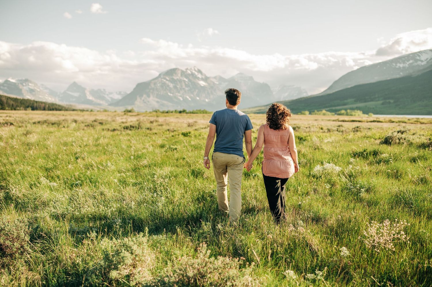 Two people walk hand in hand through a scenic mountain meadow with snow-capped peaks in the distance.