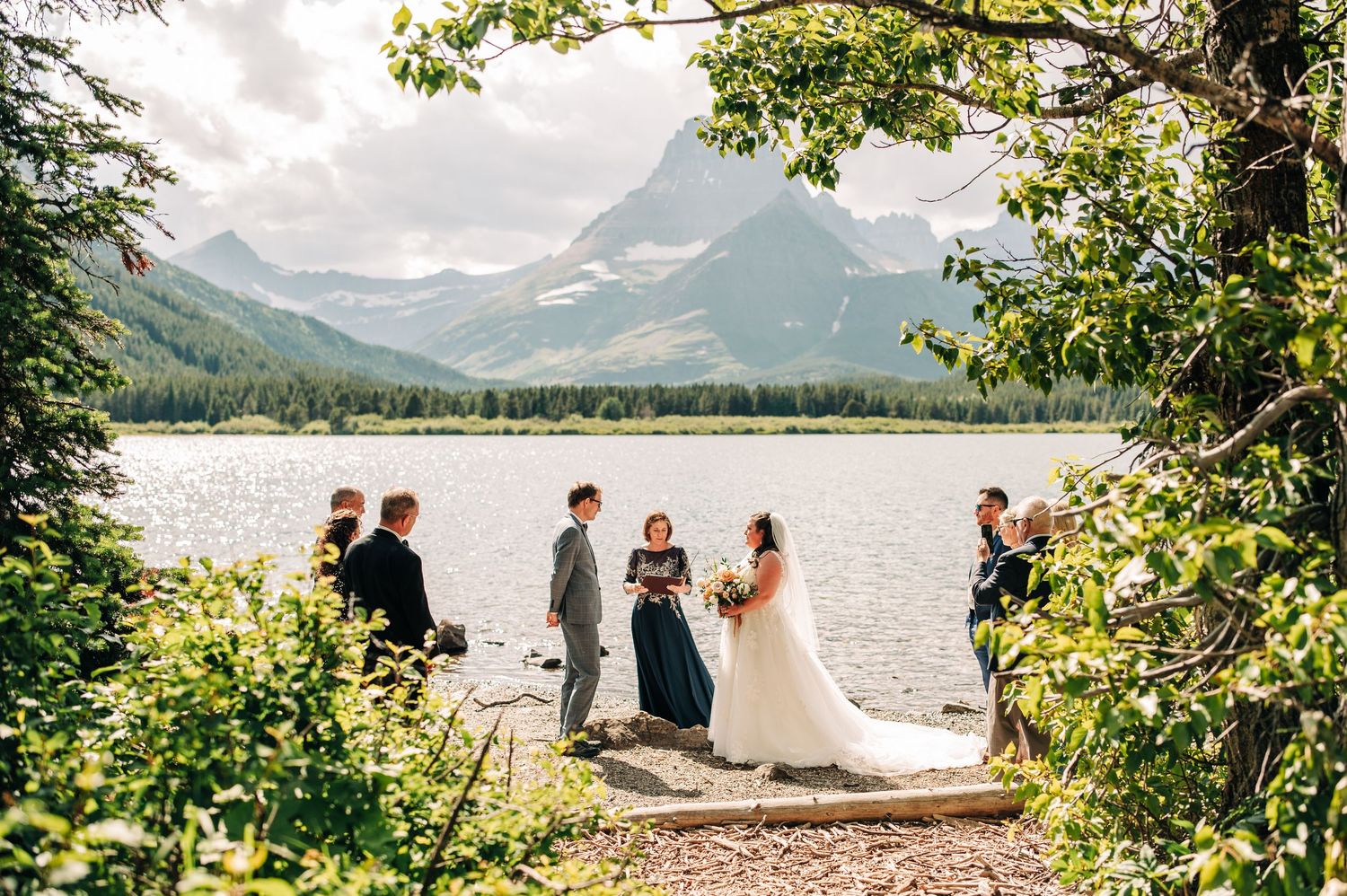 An outdoor wedding ceremony takes place on a wooden dock beside a mountain lake surrounded by evergreen trees and peaks.