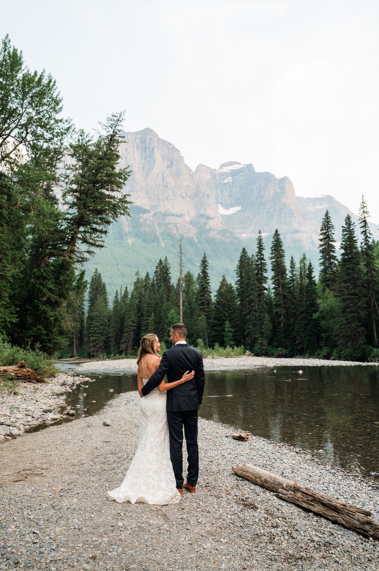 Bride and groom stand by a rocky riverbank surrounded by pine trees and a stunning mountain backdrop.