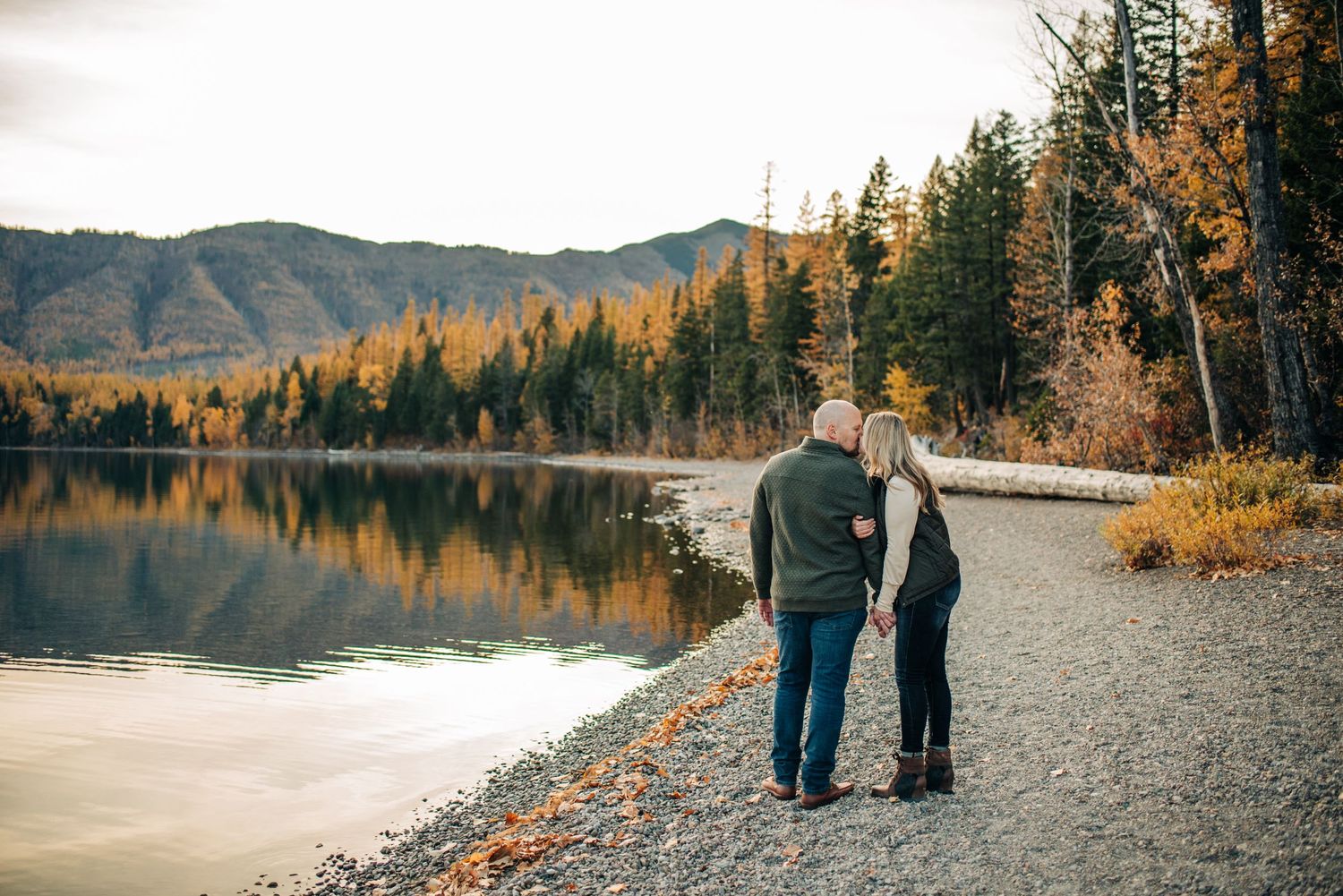 A couple walks along a lakeside path surrounded by autumn trees and mountains reflected in calm waters.