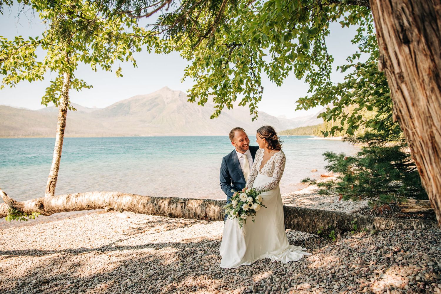 A couple poses together on a scenic lakeshore with mountains and trees framing the romantic waterfront setting.