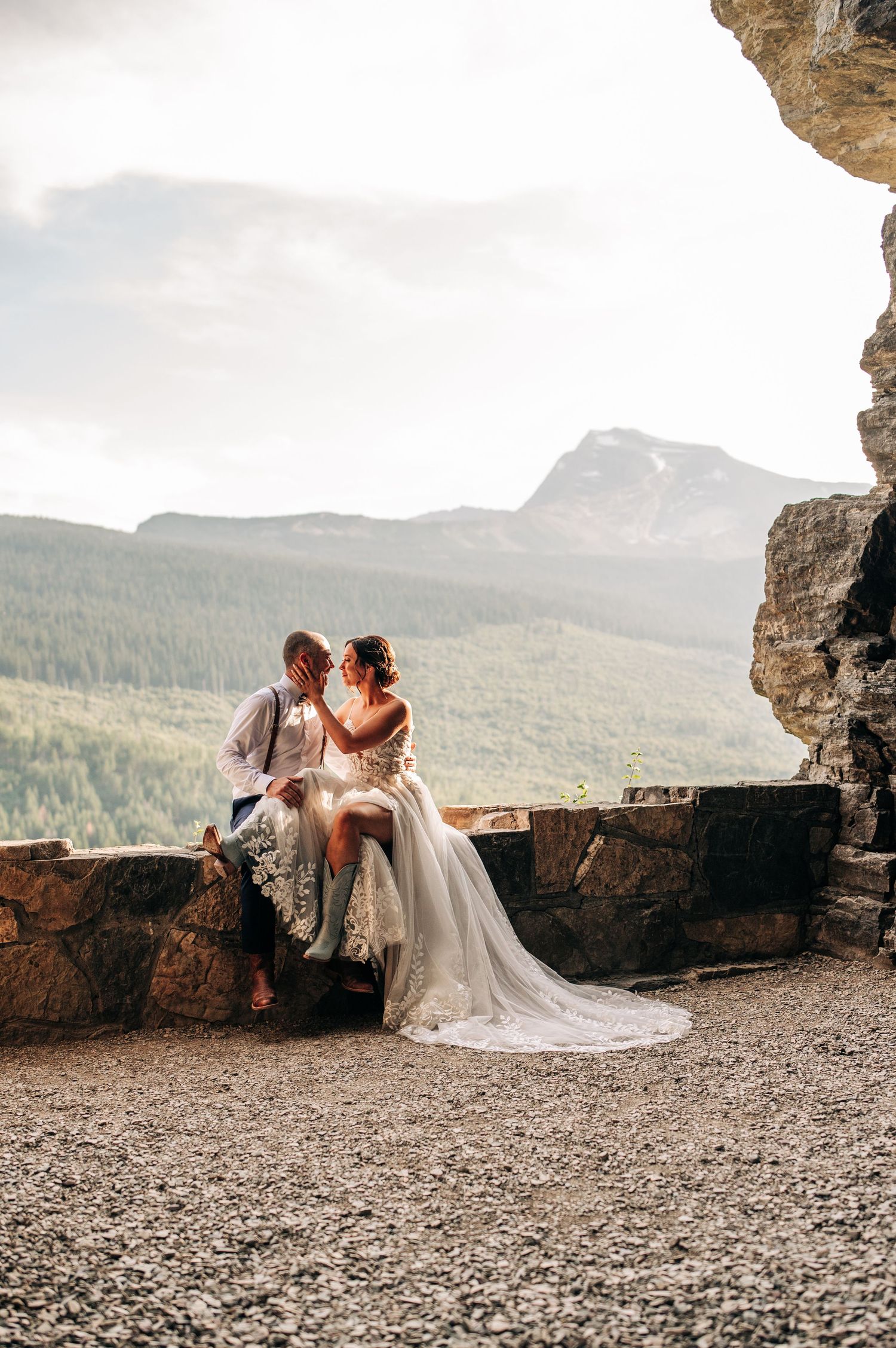 A couple in wedding attire sits at a scenic mountain overlook with dramatic valley views and rocky terrain.