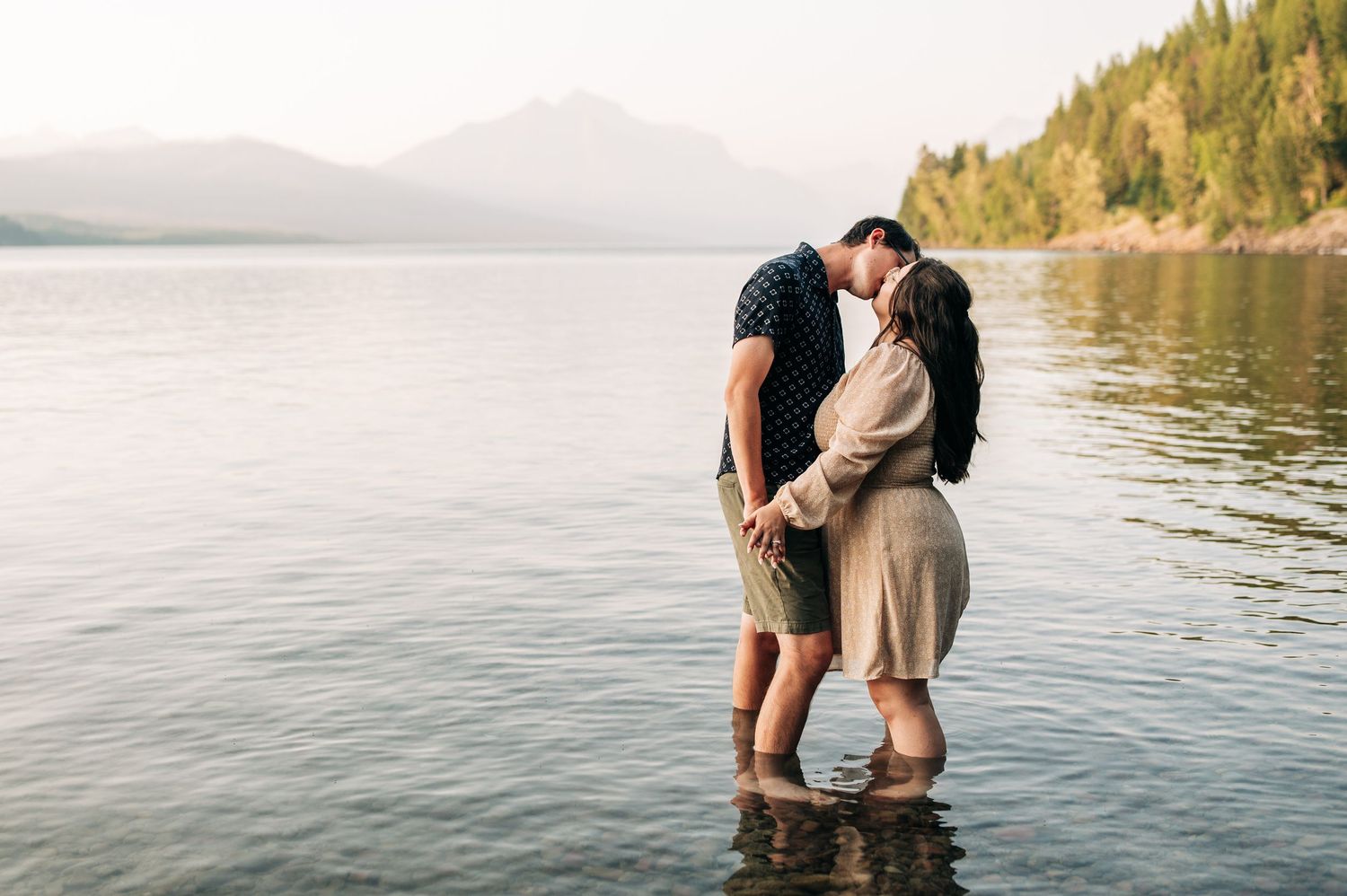 A couple shares an intimate moment while standing in shallow lake water with mountains and forest in the background.