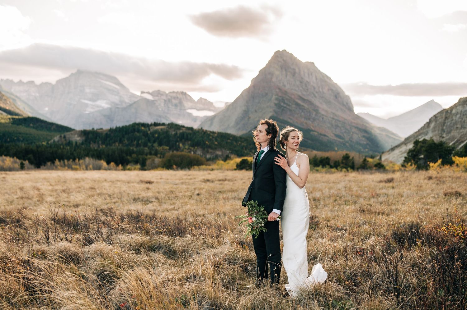 A couple in wedding attire stands in tall golden grass with majestic mountain peaks in the background at sunset.