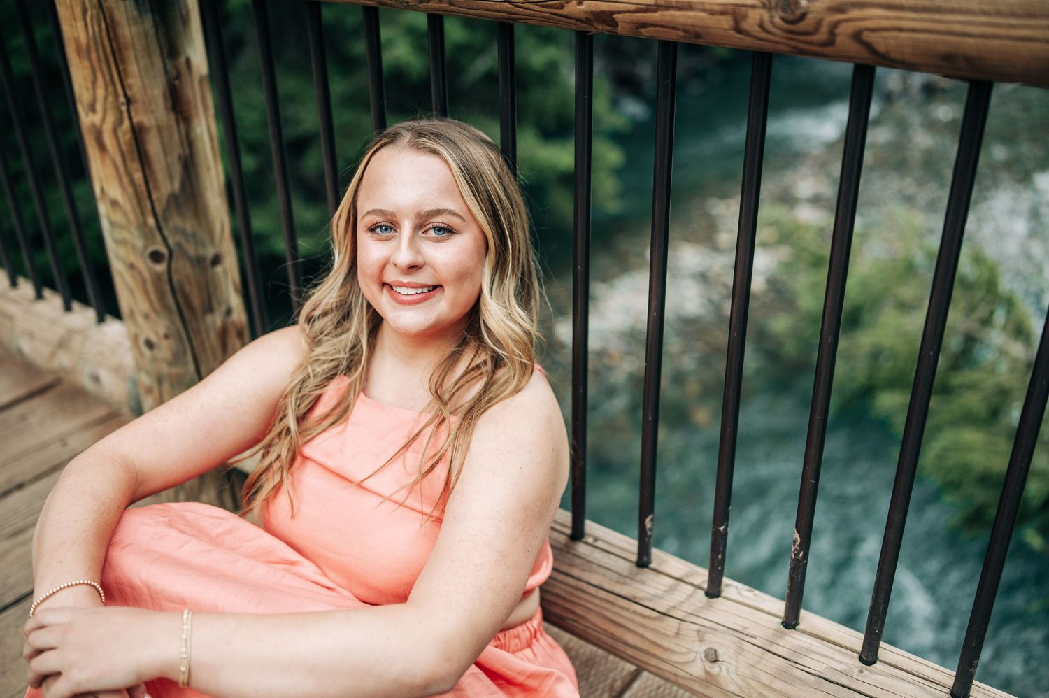 A person in a coral dress smiles warmly while sitting on a wooden deck with green railings and water in the background.